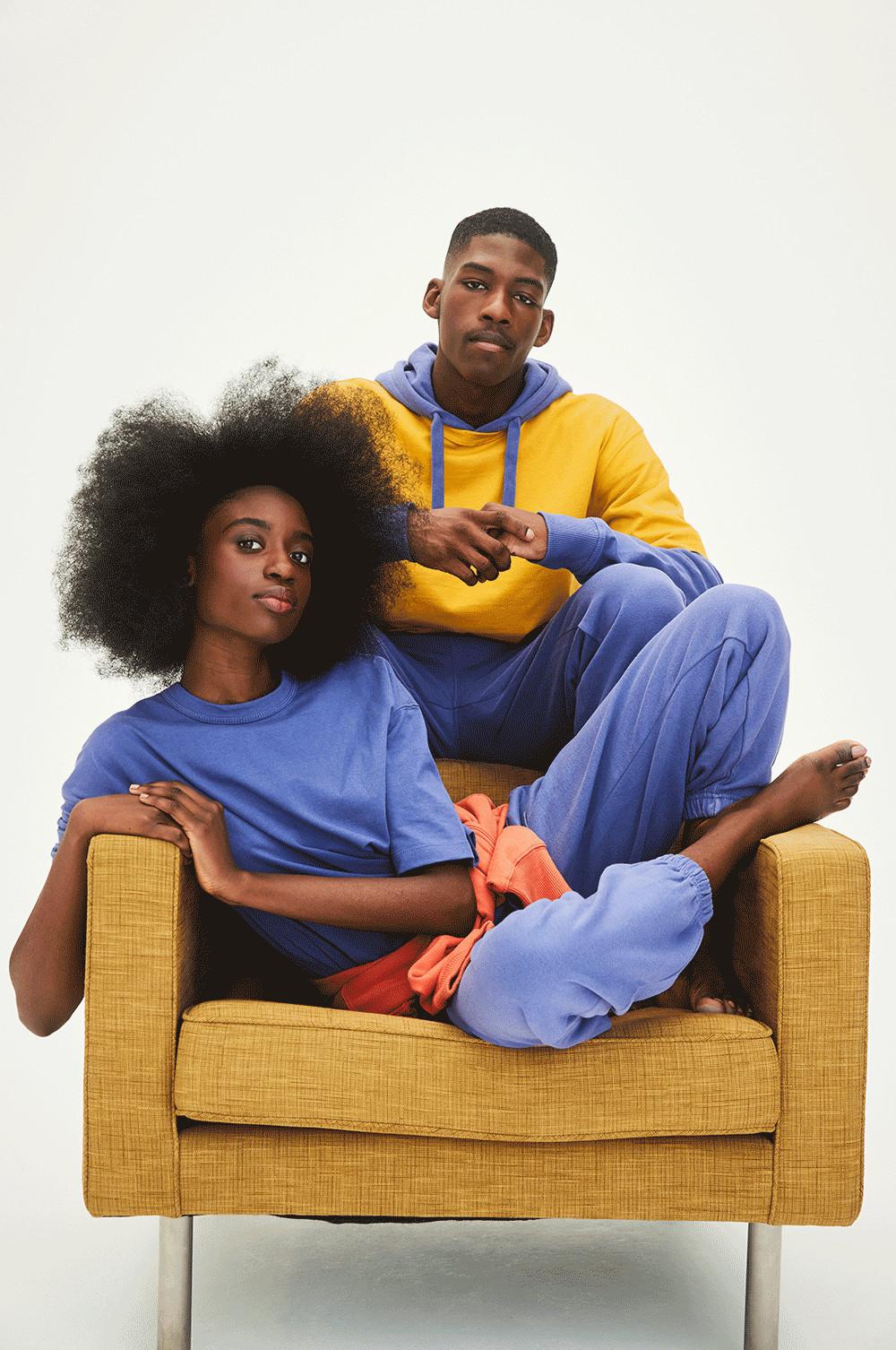 Models sit in mustard chair, woman wears purple set and man in yellow