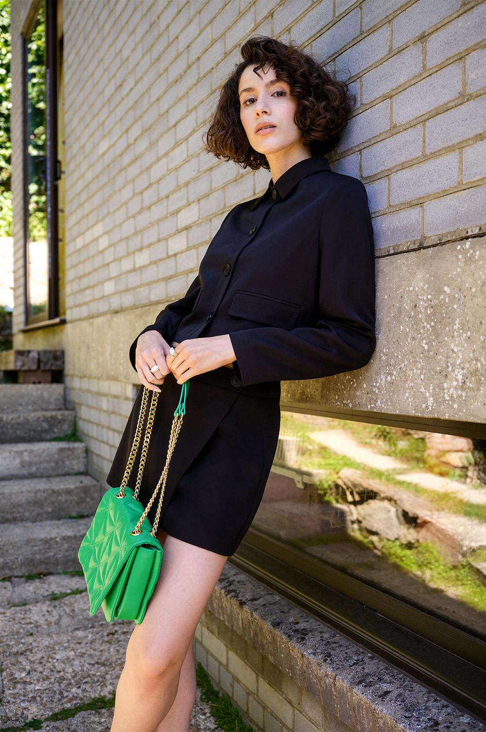 Model wears black cropped blazer and mini skort coord with green accessories