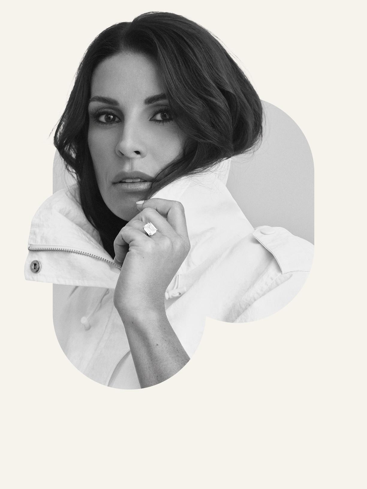 Person wearing mauve sports bra with black sweater raised above head, against pink background with white oval shape.