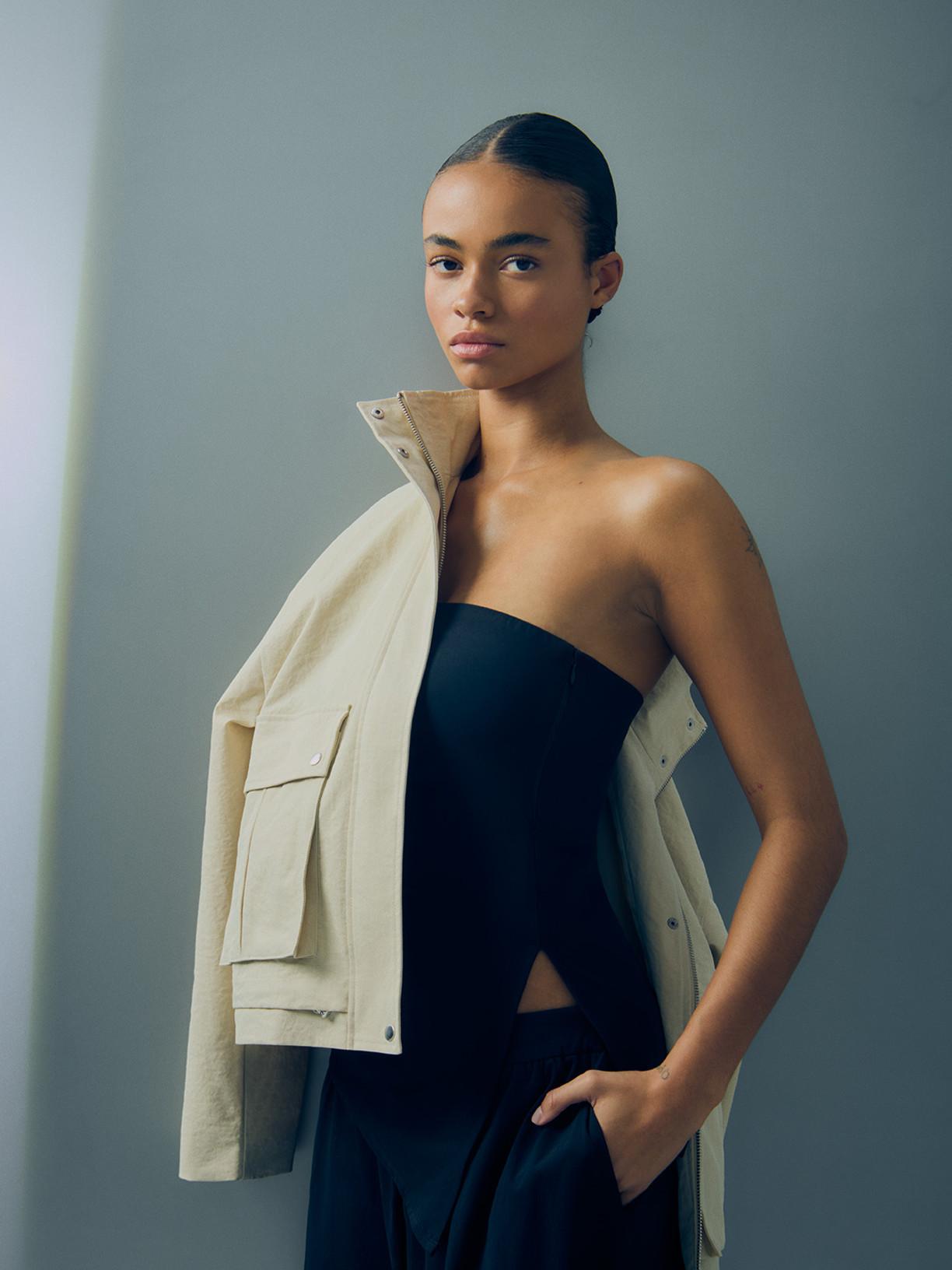 Model wearing black strapless jumpsuit with beige utility jacket draped over shoulder.