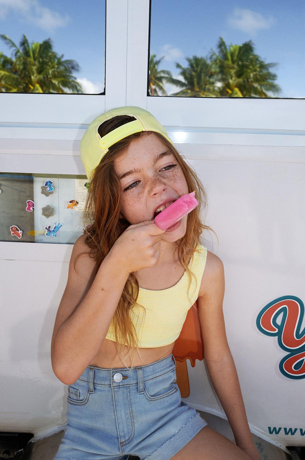 Child with ice lolly wearing denim shorts, yellow cropped swim top and yellow cap