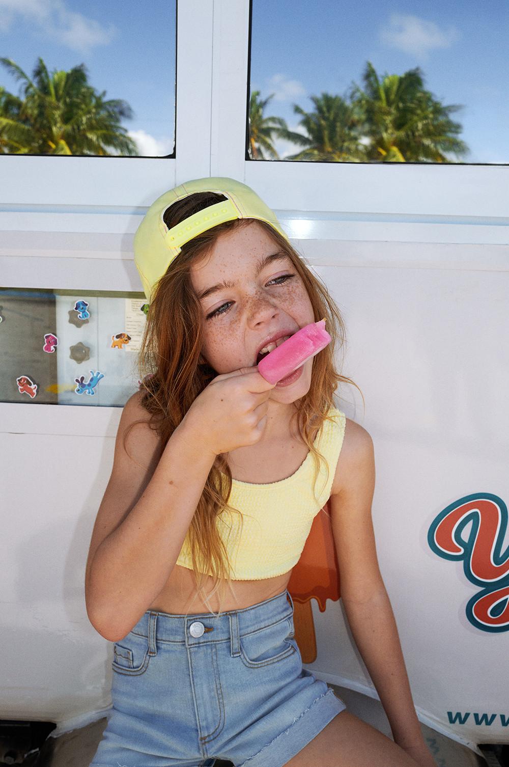 Modelo infantil con pantalones vaqueros cortos, gorra amarilla y top de baño corto amarillo