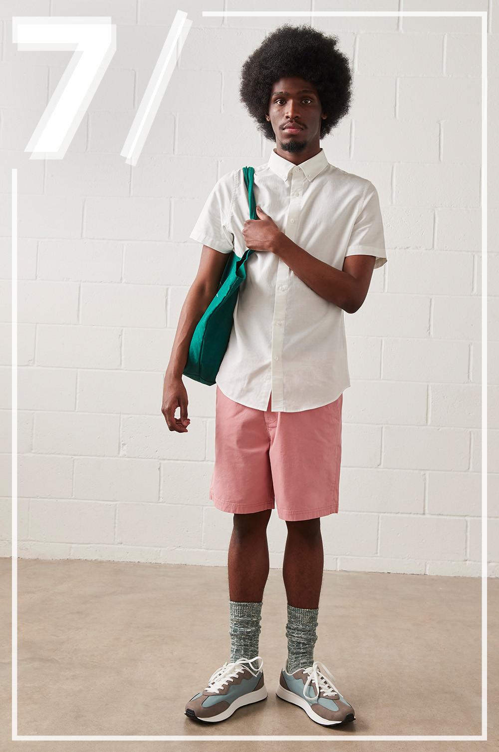 model wears white short sleeved oxford shirt, with pink shorts and green bag