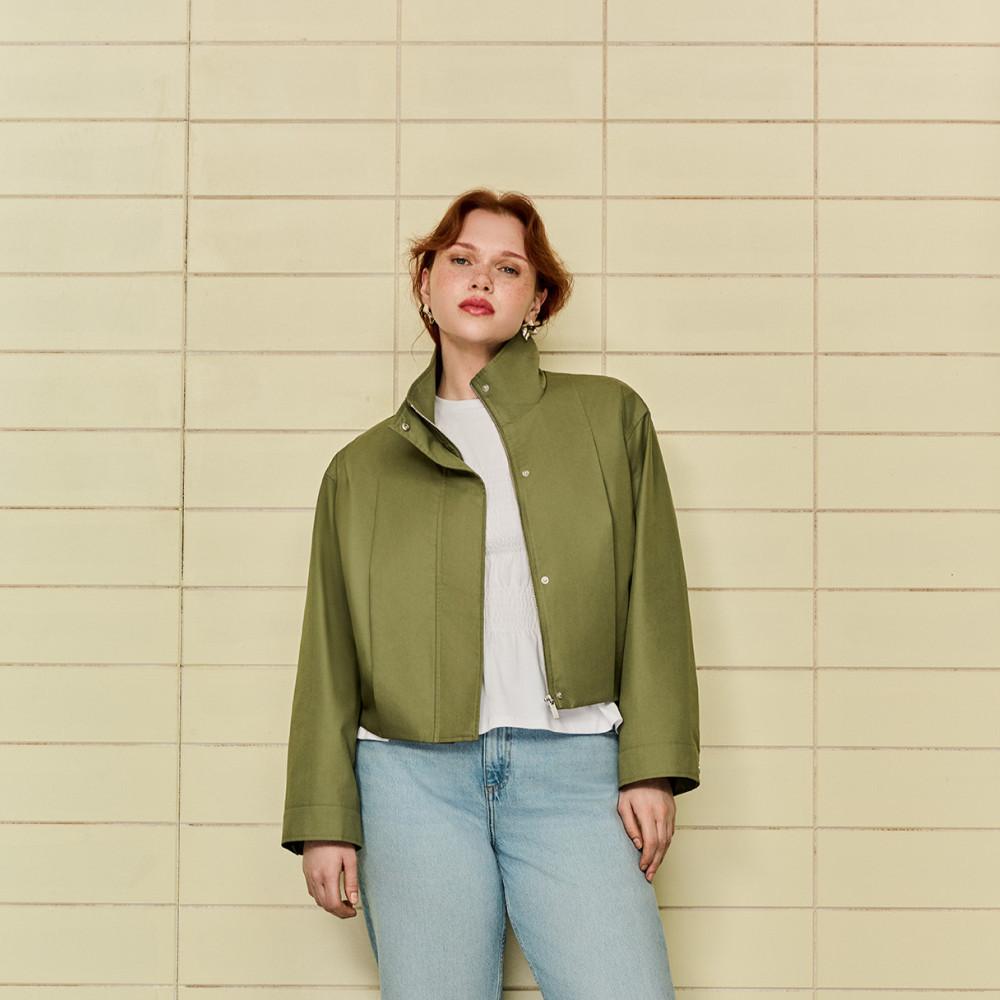 Kort olijfgroen jack met wit topje en lichte spijkerbroek met een betegelde muur als achtergrond.