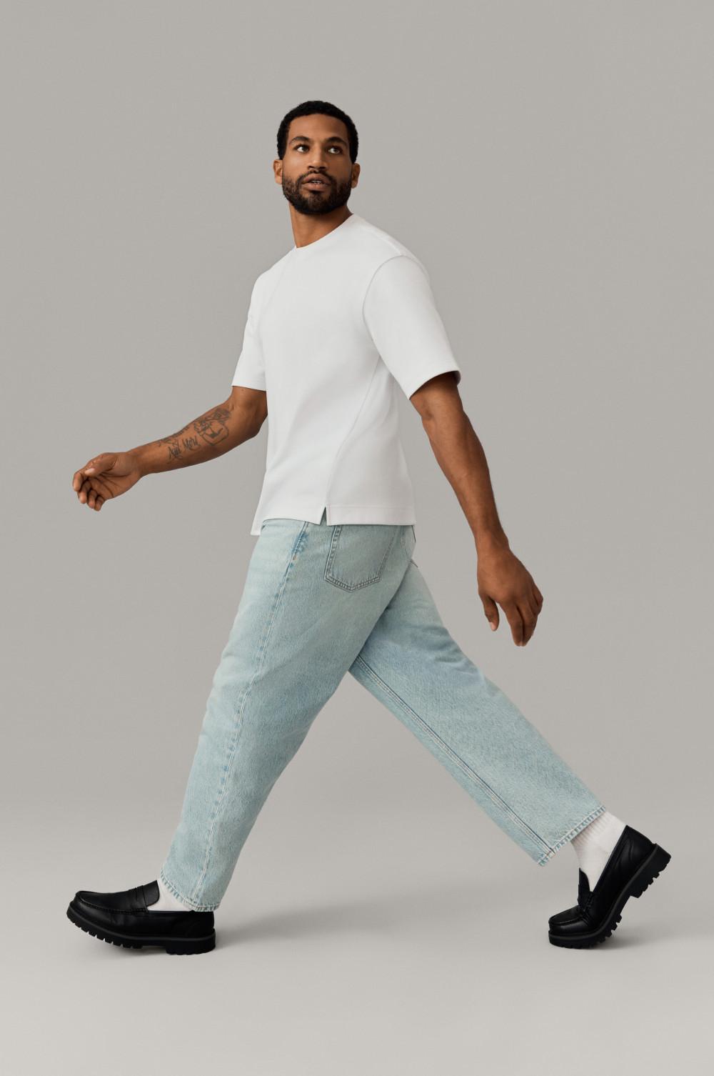 Person walking in white short-sleeve top, light blue relaxed jeans and black loafers against neutral backdrop.
