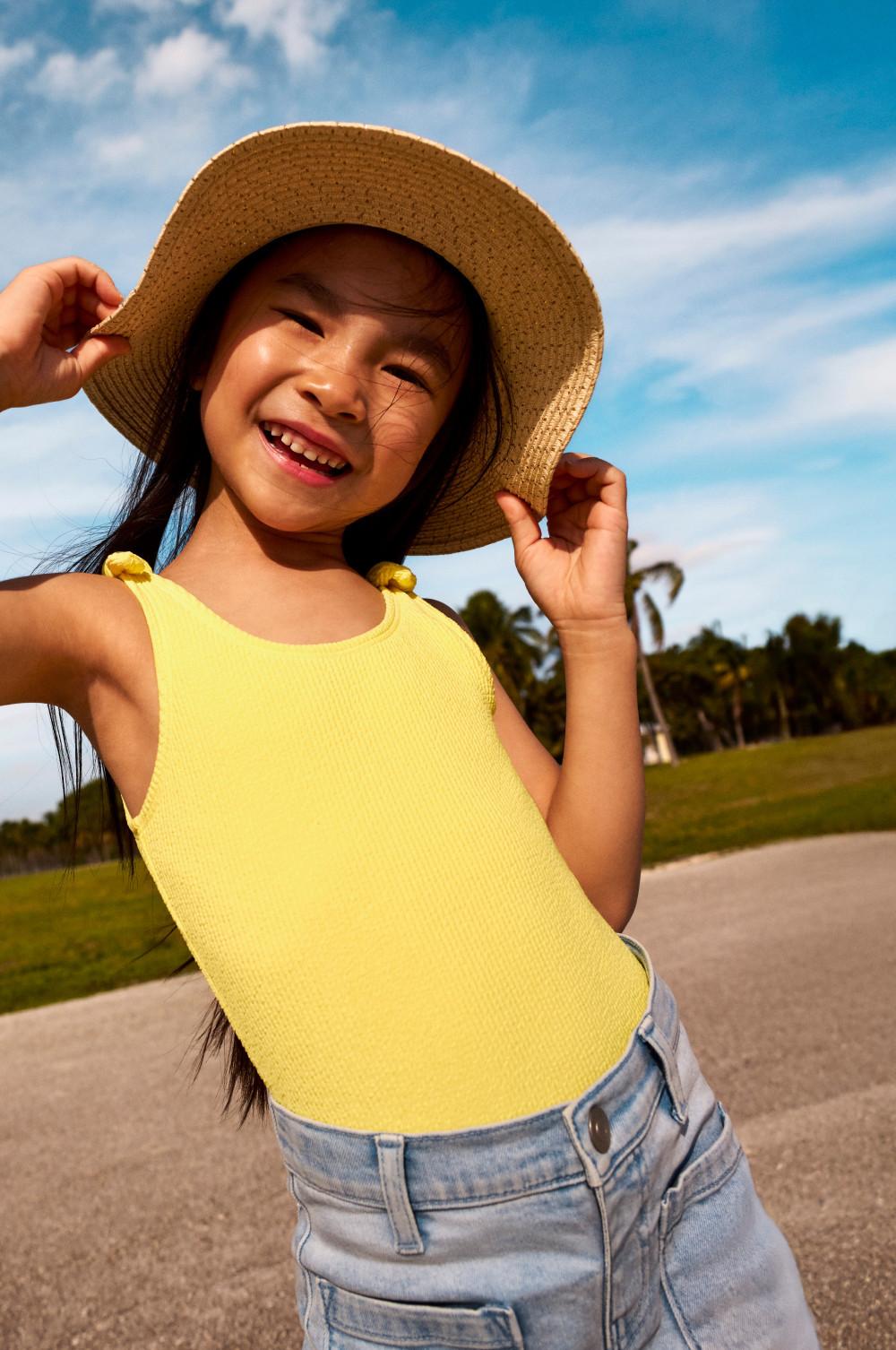 Young girl smiling outdoors wearing a yellow vest top and straw sun hat with light blue jeans.