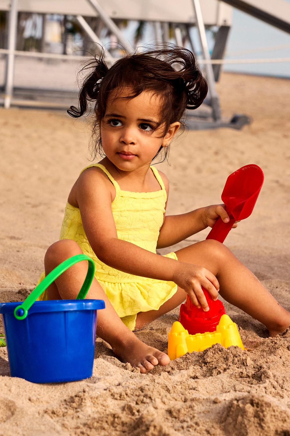 Young girl in yellow swimsuit playing with colourful beach toys including bucket and spade on sandy beach.
