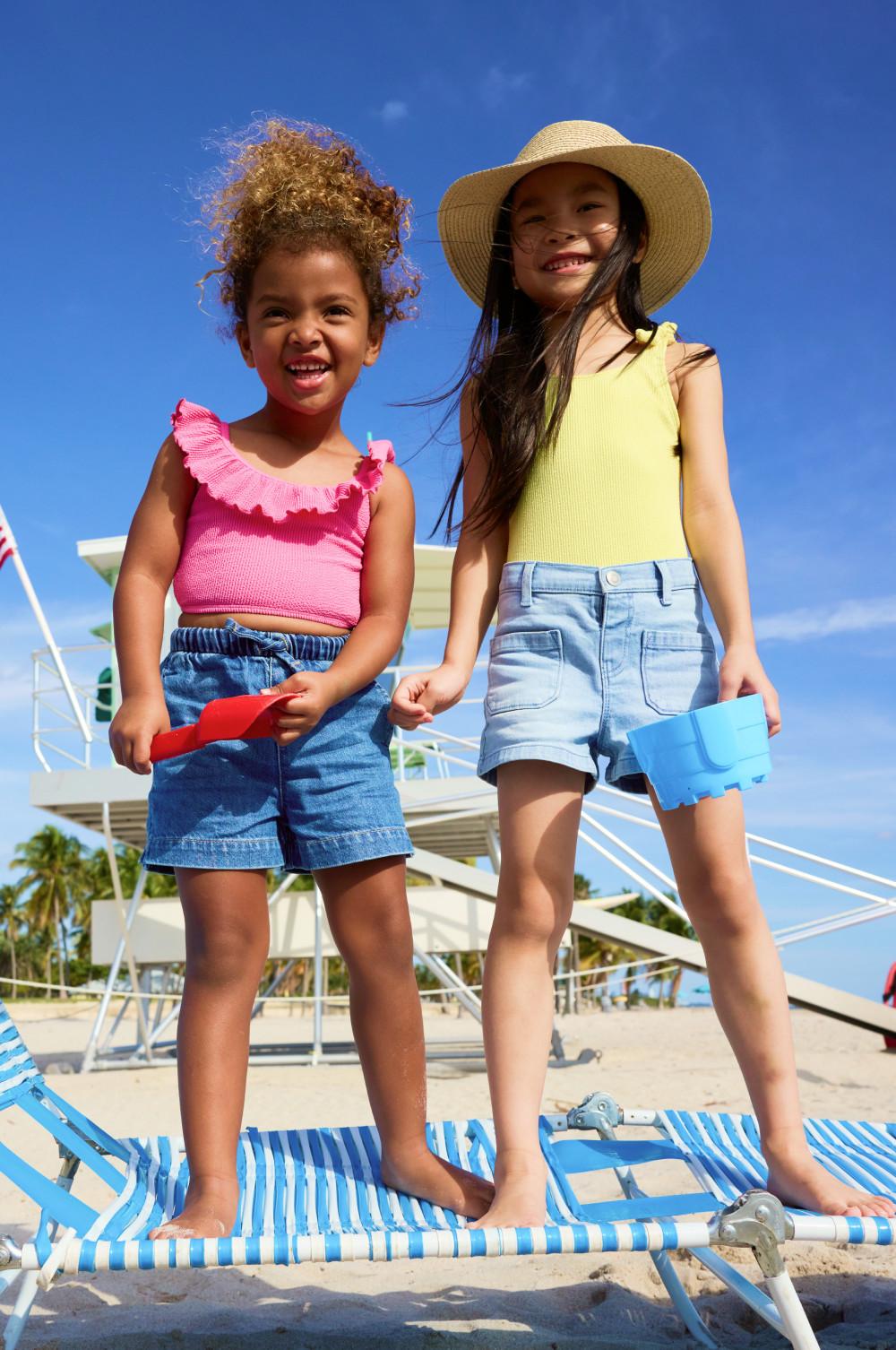 Two girls standing on poolside loungers wearing summer outfits with denim shorts and colourful tops.