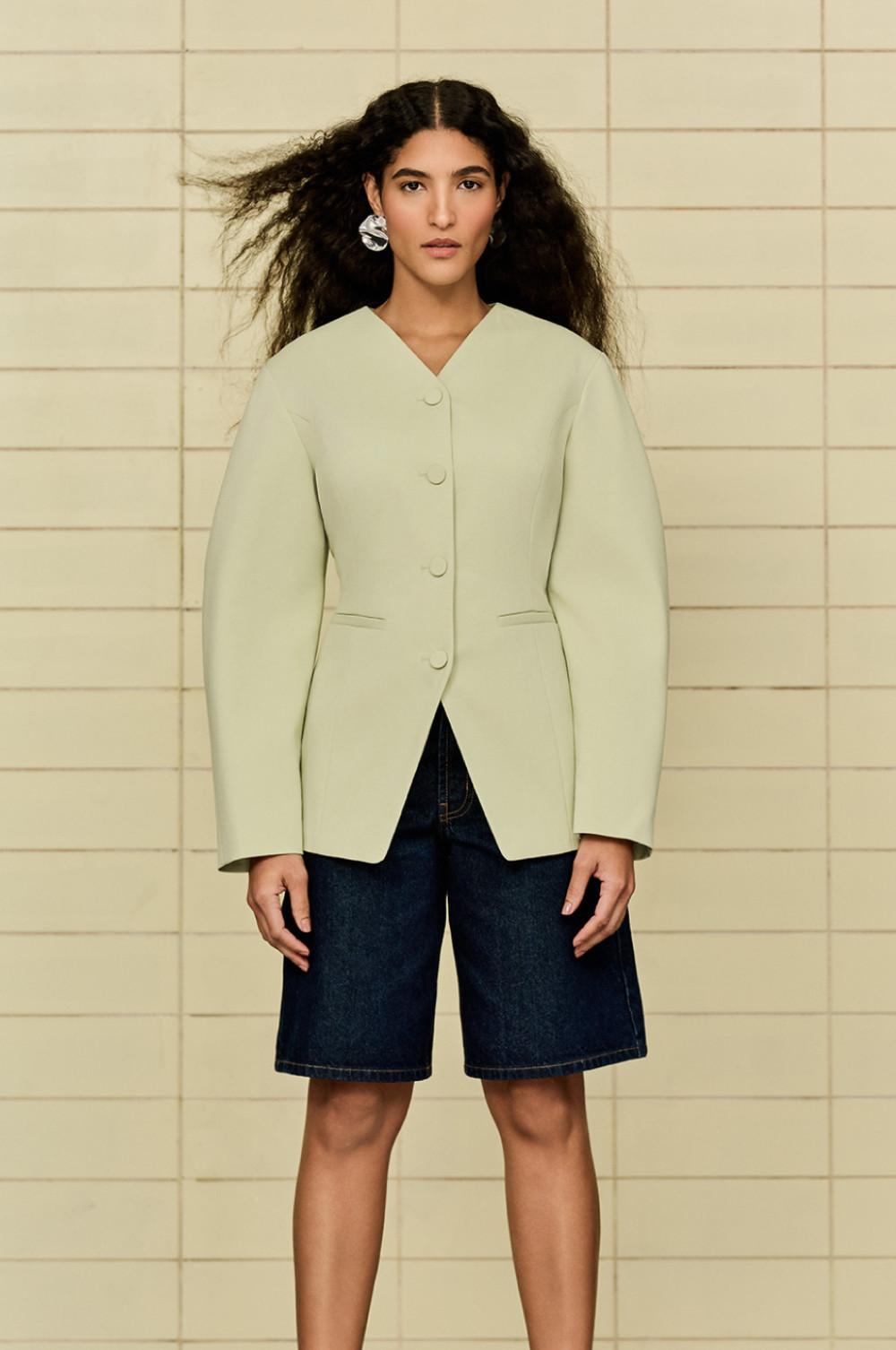 Een model staat voor een tegelwand en draagt een licht mintkleurige blazer met textuur en knoopsluitng voor op een donkere denim bermuda.