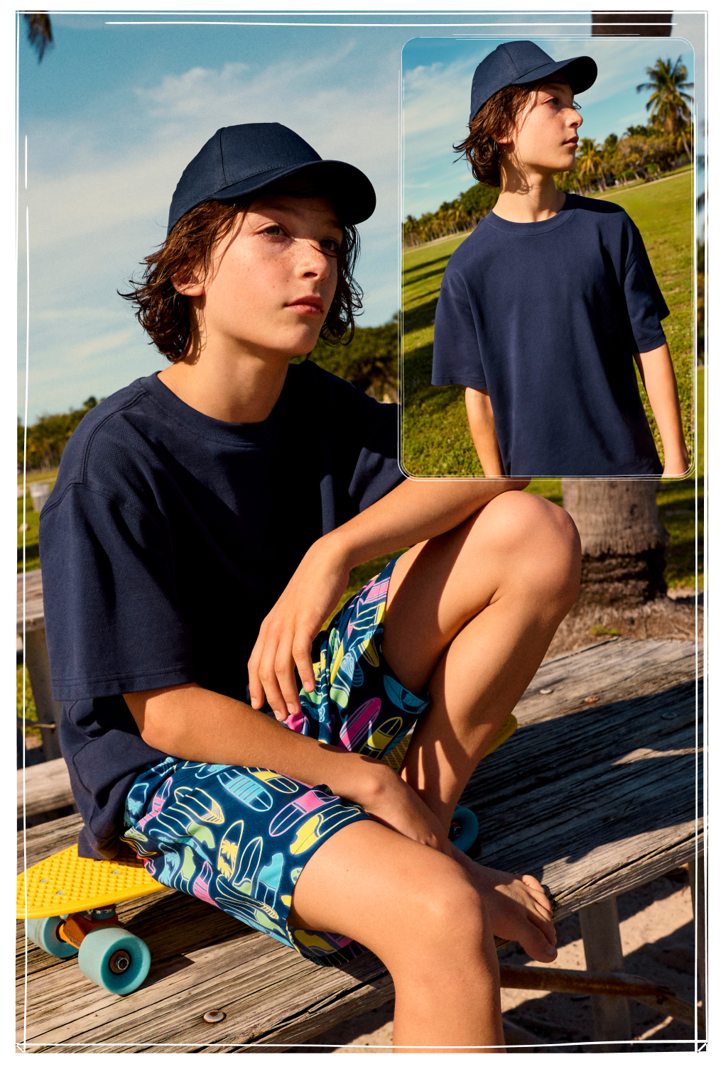 Boy wearing navy baseball cap and t-shirt with colourful printed shorts sitting on skateboard outdoors.