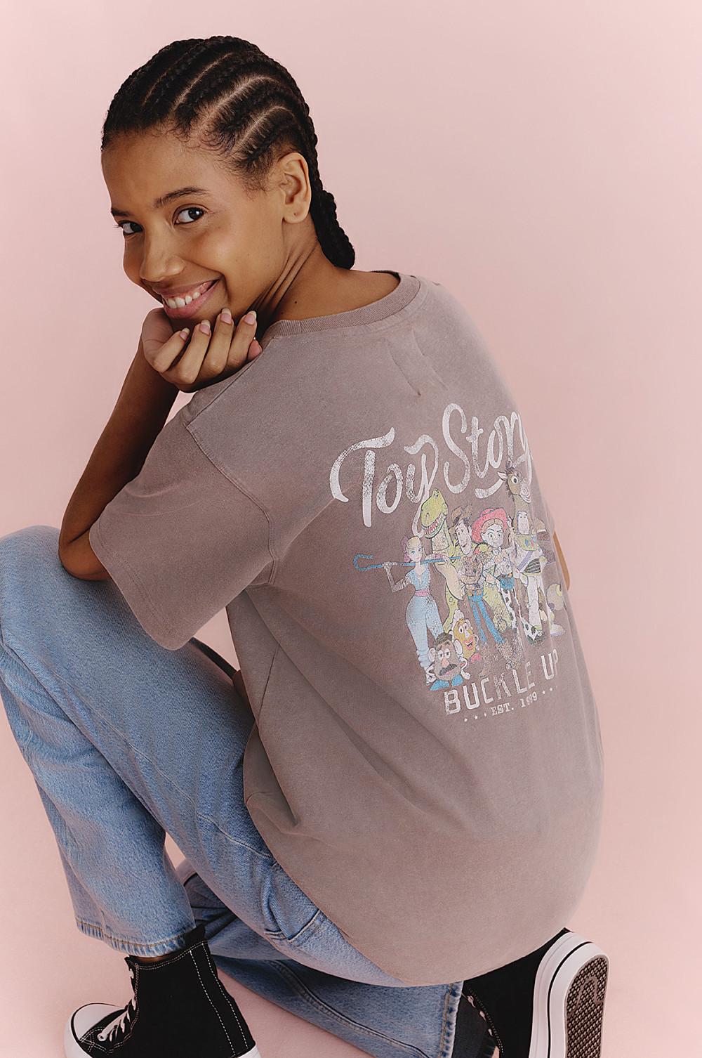 Model wearing a brown Toy Story graphic sweatshirt with Toy Story character print, sitting and smiling.