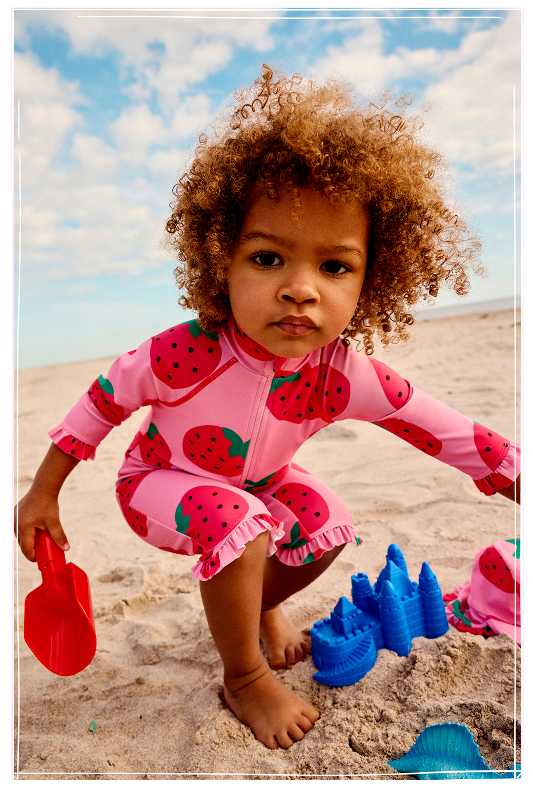Young girl smiling on beach chair wearing pink heart-shaped sunglasses, pink ruffled swimsuit and denim shorts.