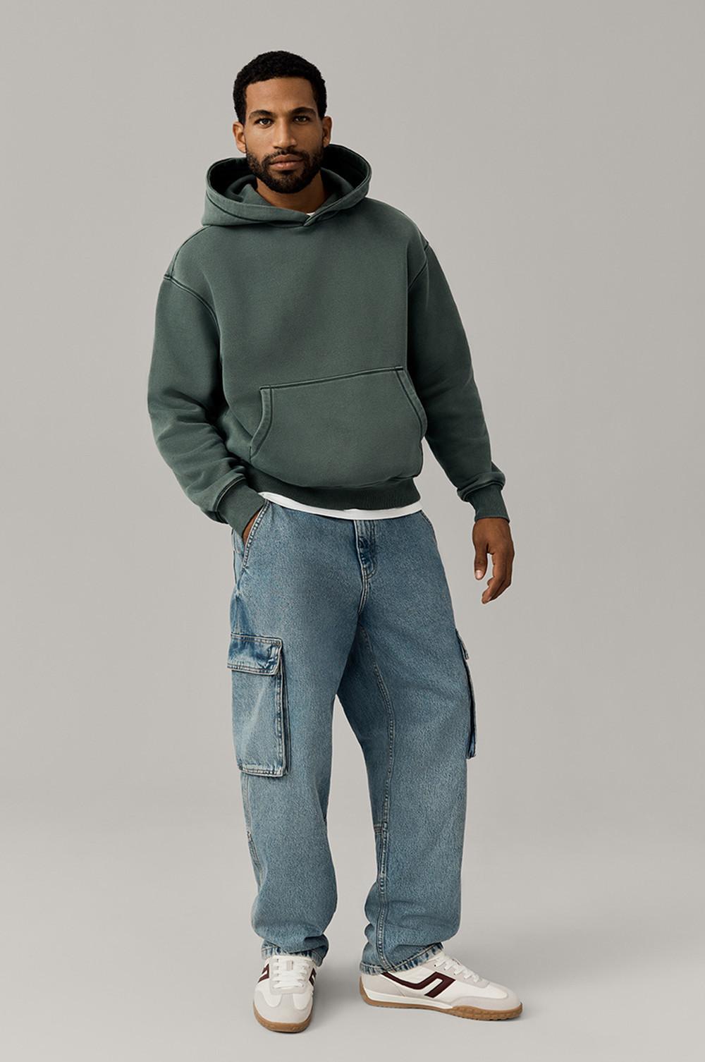 Person wearing green hoodie with blue cargo jeans and white sneakers against neutral background.
