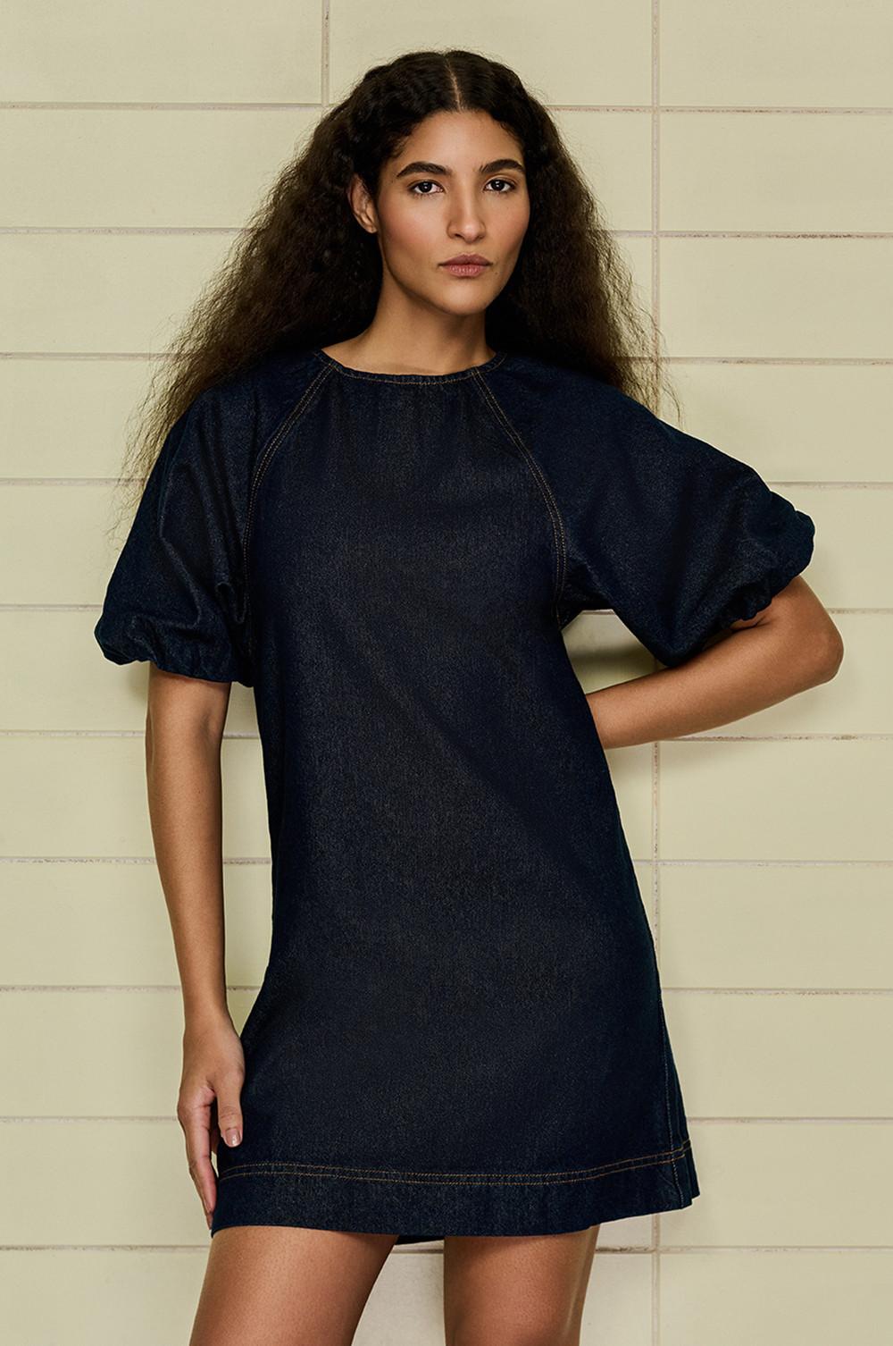 Donkere mini-spijkerjurk met pofmouwtjes en contrasterende stiksels met een tegelwand op de achtergrond.