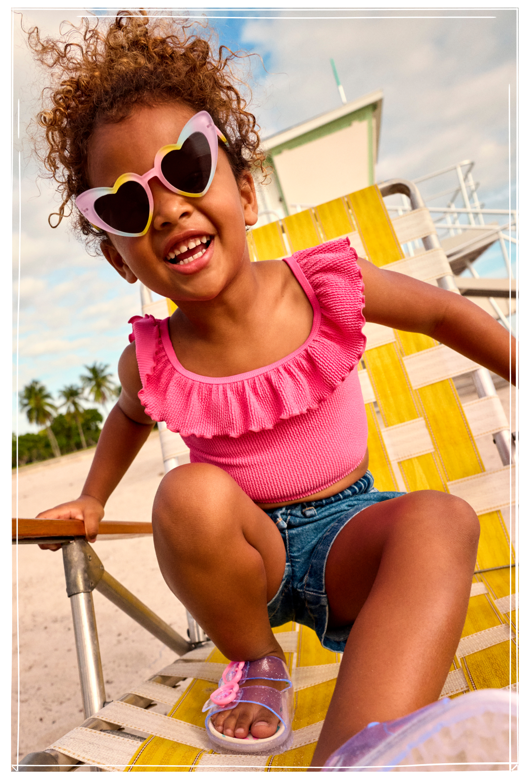 Young girl smiling on beach chair wearing pink heart-shaped sunglasses, pink ruffled swimsuit and denim shorts.