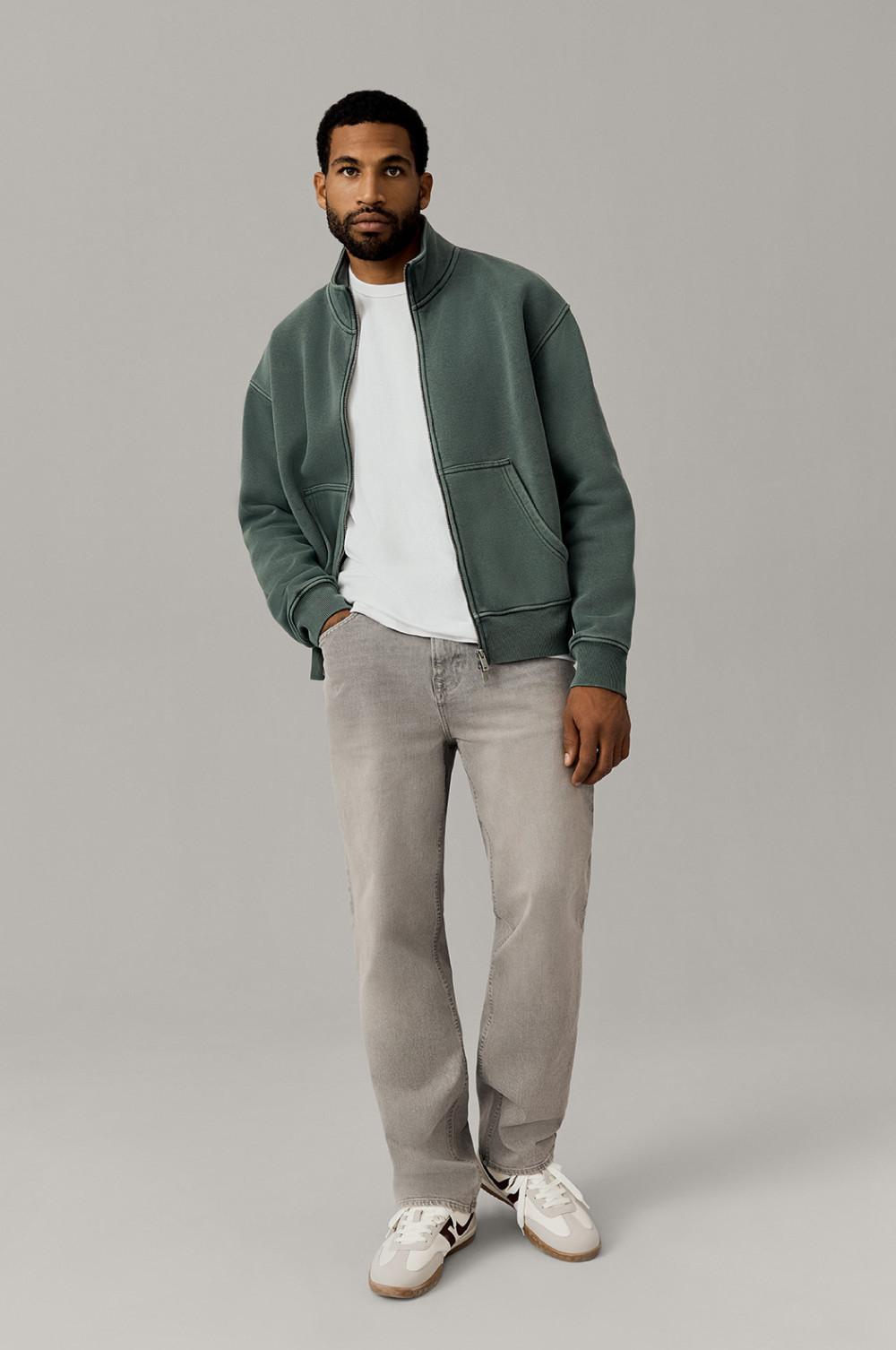 Person wearing green zip-up jacket over white T-shirt with light gray jeans and white sneakers.