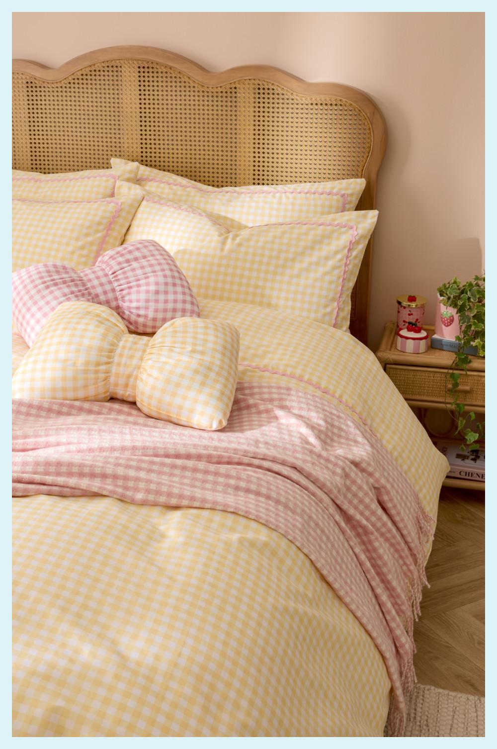 Bed with yellow gingham duvet, pink throw, decorative cushions and rattan headboard beside small nightstand.