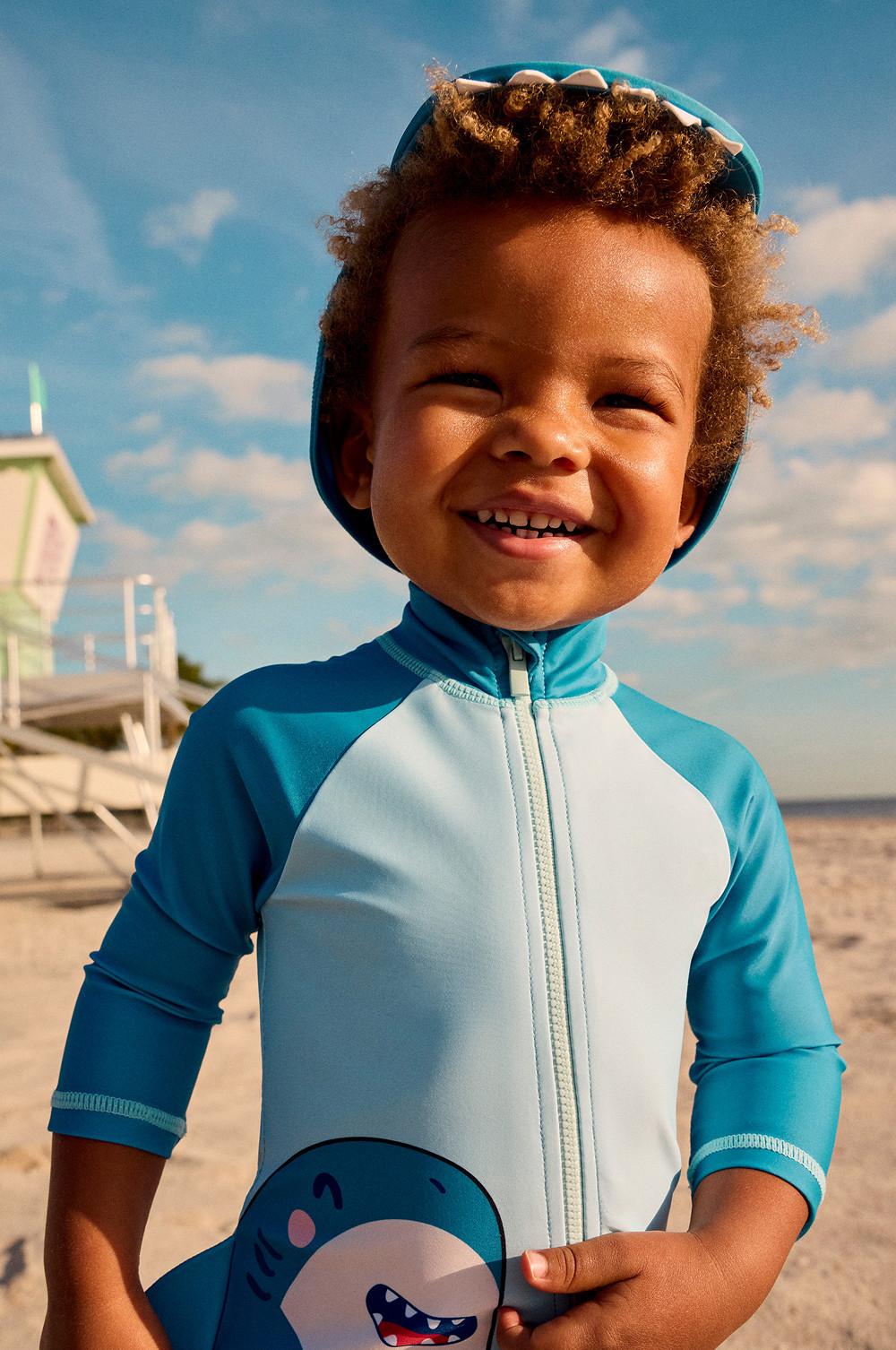 Child wearing teal and white zip-up rash vest with dinosaur design and matching swim hat at the beach.