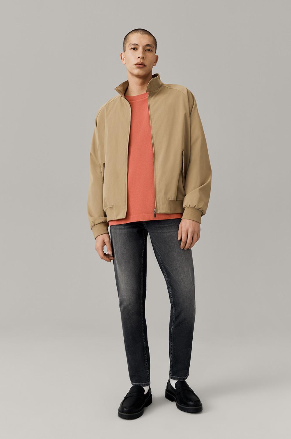 Person wearing beige bomber jacket over coral T-shirt, paired with gray jeans and black loafers.