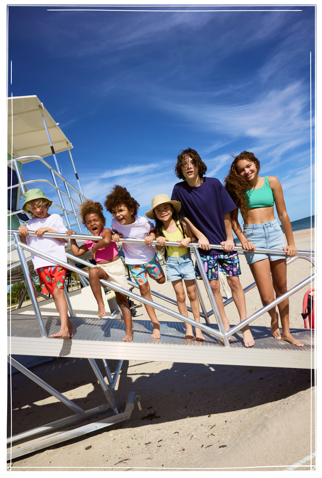 Seven children standing on a lifeguard tower at the beach wearing colourful summer clothing and swimwear.