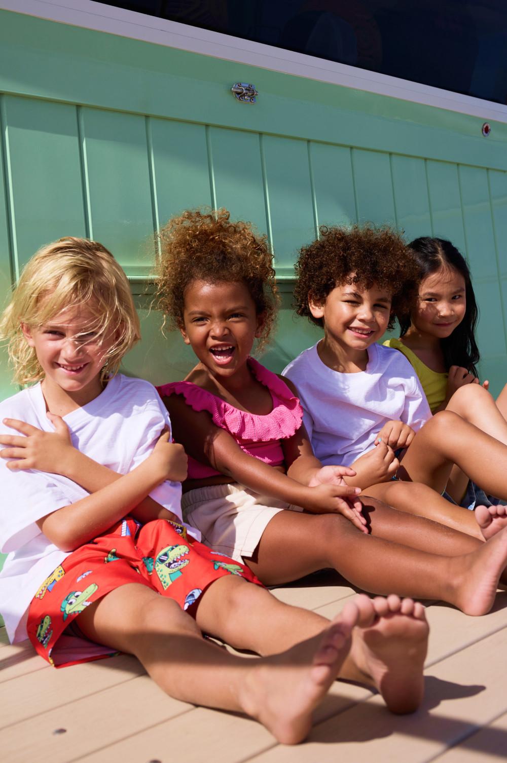 Four children sitting together by a pool wearing colourful swimwear and smiling at the camera.