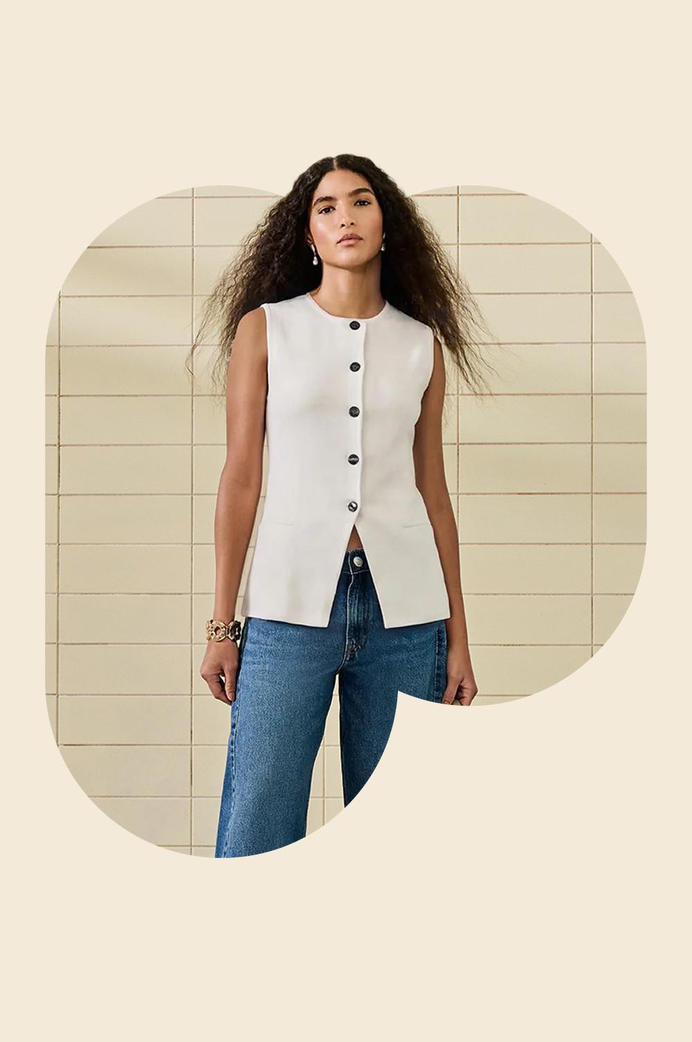 Wit mouwloos gilet met knoopsluiting met een blauwe spijkerbroek met een tegelwand als achtergrond.