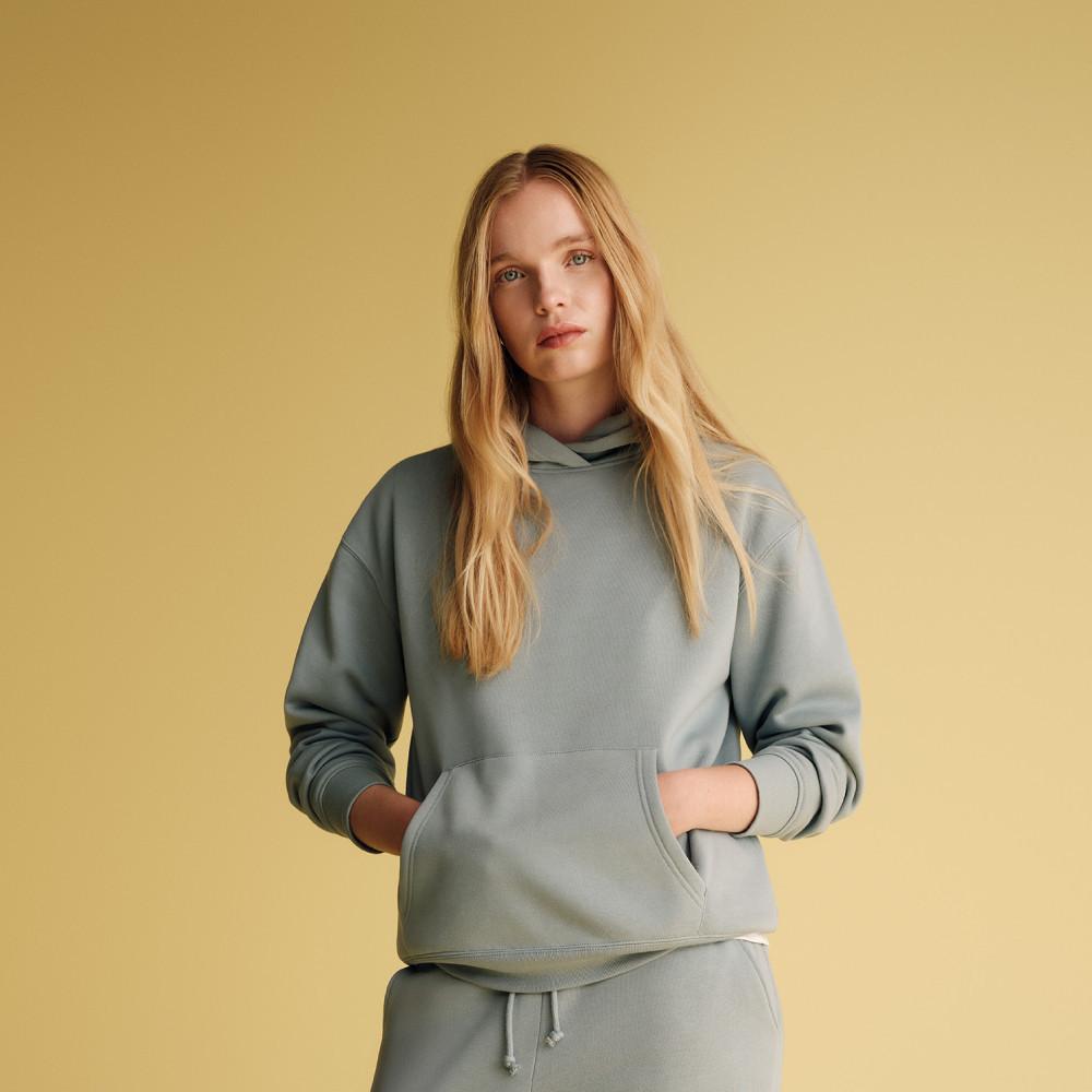 Persona con las manos en el bolsillo canguro de una sudadera en verde salvia con capucha y joggers a juego sobre un fondo beis cálido.