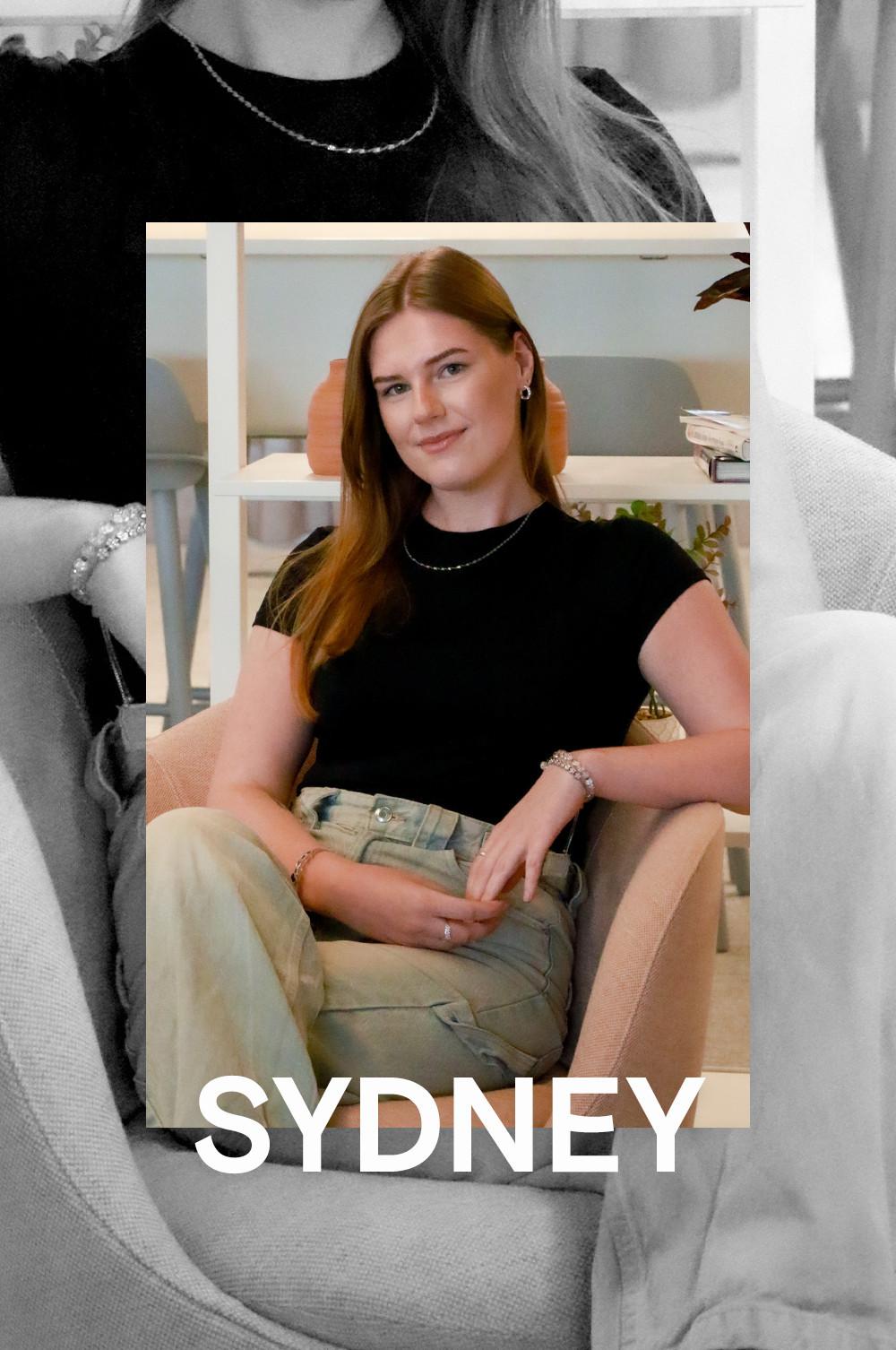 Sydney Lasek draagt ​​een zwart topje met korte mouwen, een subtiele ketting en een lichte spijkerbroek.