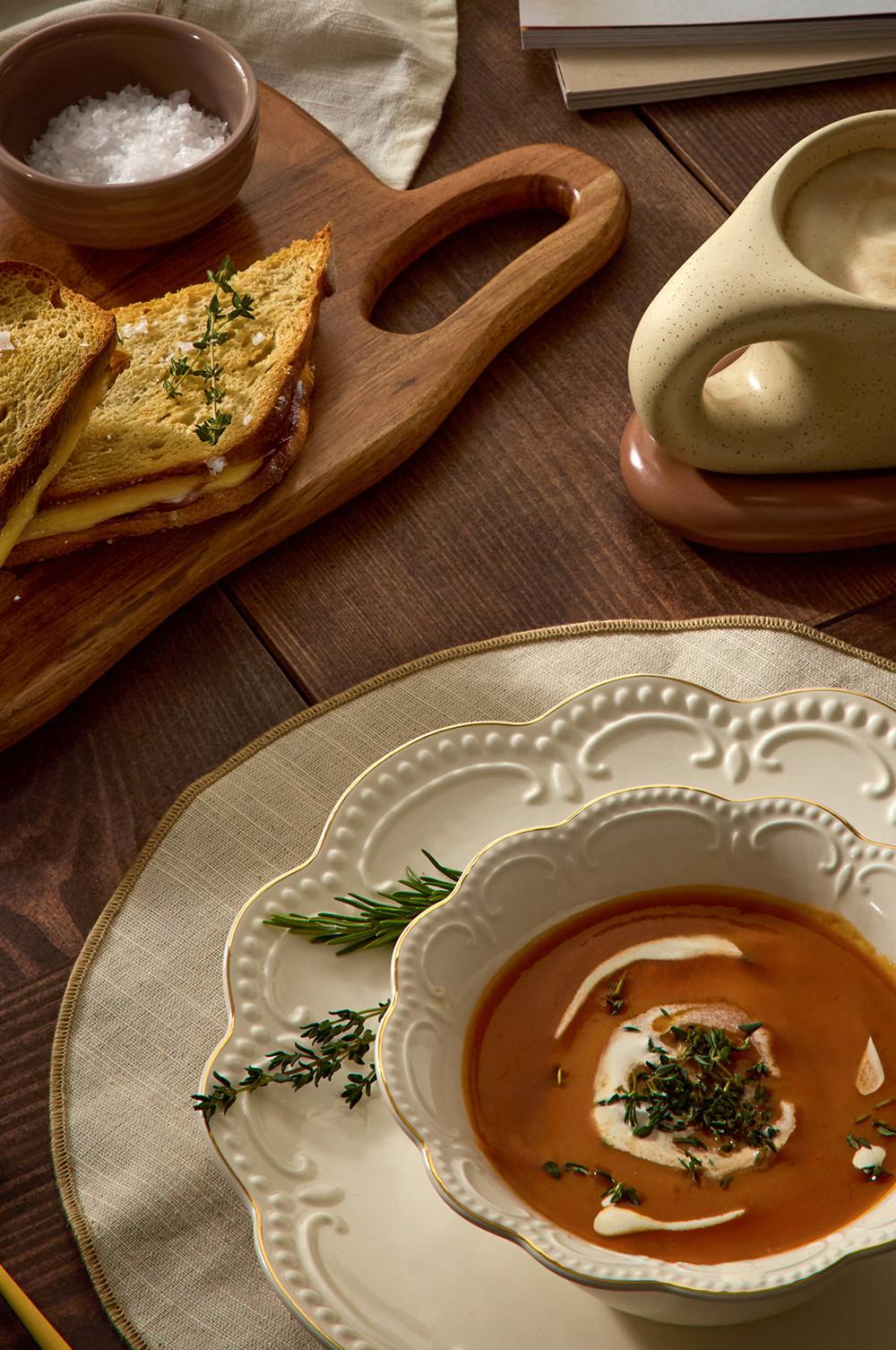 Creamy soup with herb garnish in decorative bowl, served with grilled cheese sandwich and salt on wooden board.