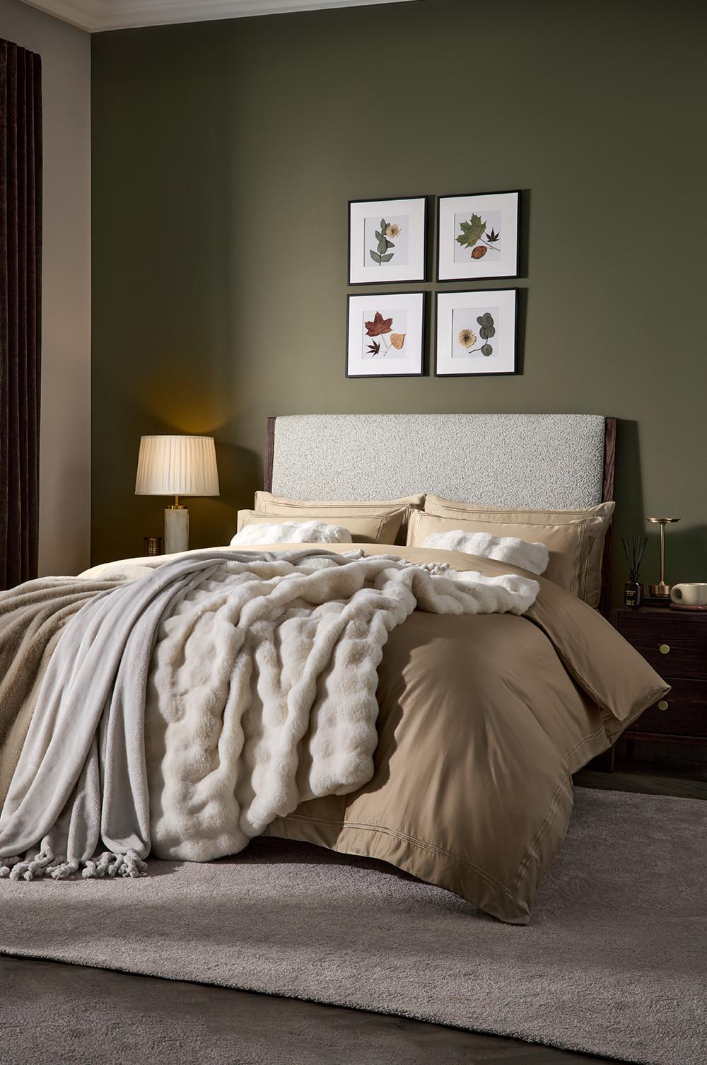 Cosy bedroom with beige bedding, plush throw blanket, upholstered headboard and botanical prints on dark green wall.