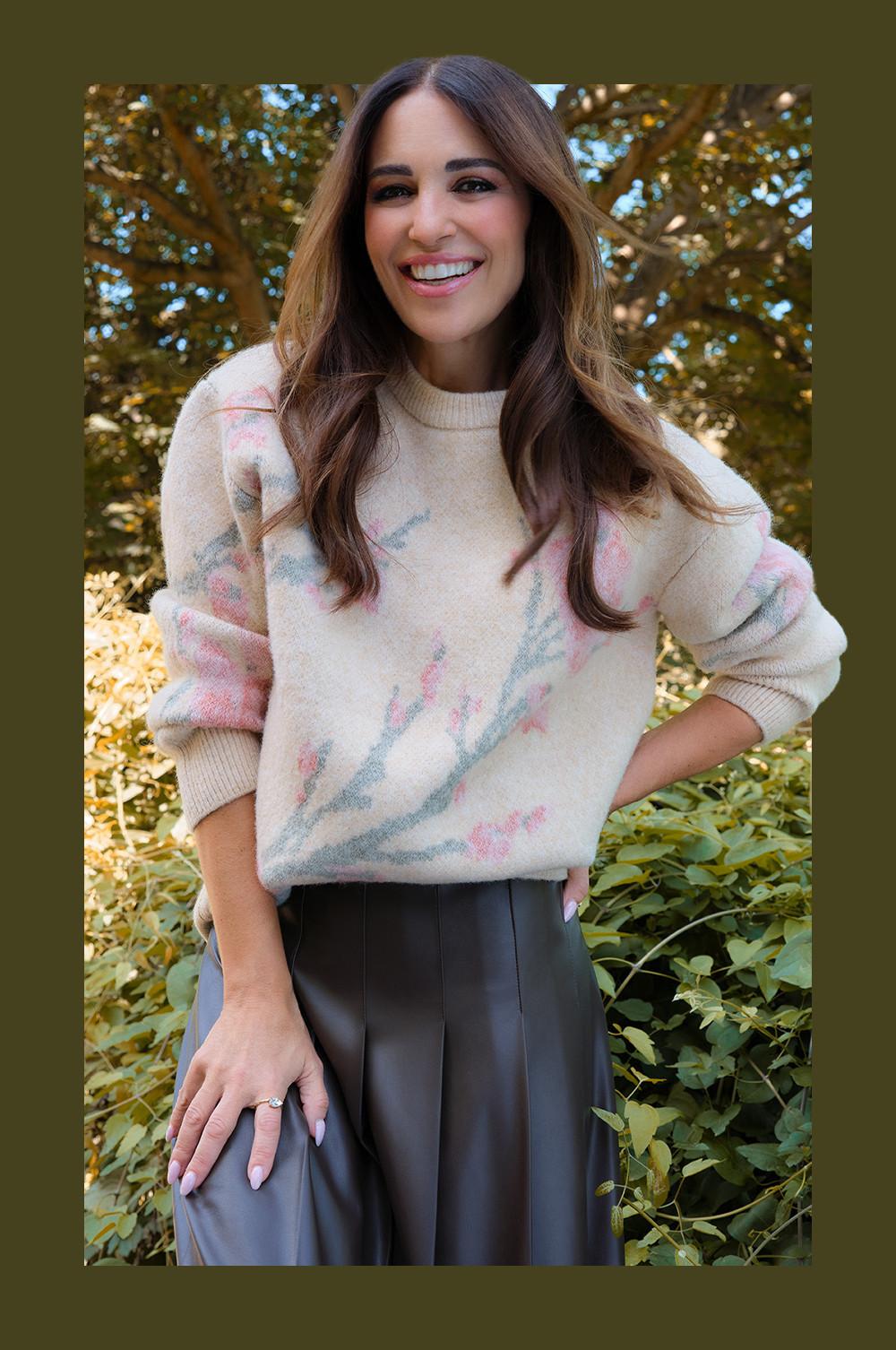 Pull crème à motif rose et bleu porté avec un pantalon gris foncé, avec un jardin en fond.