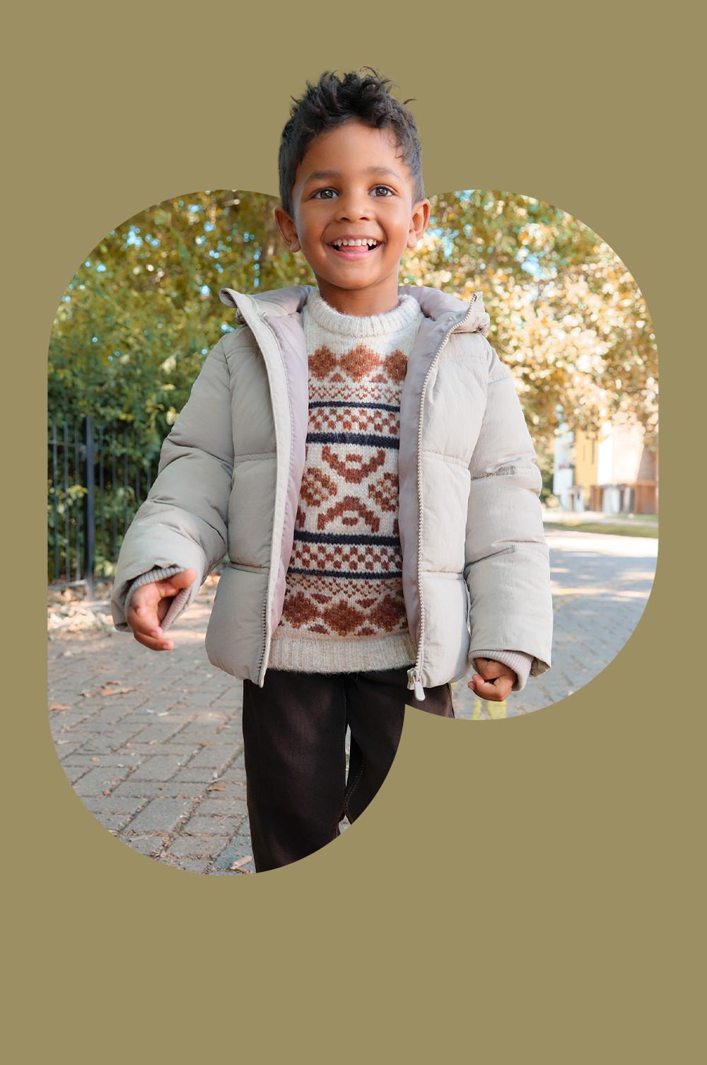 Enfant en plein air dans un décor automnal portant une doudoune beige sur un pull en tricot à motifs marron et bleu marine.