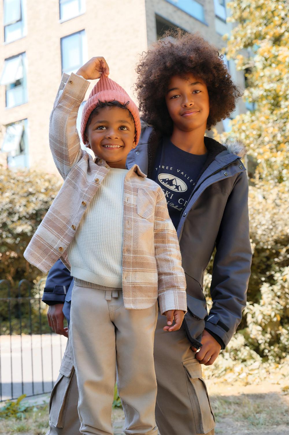 Deux enfants portant des tenues de saison : l’un avec une veste polaire marron et un pantalon rose, et l’autre une parka orange avec un jean.
