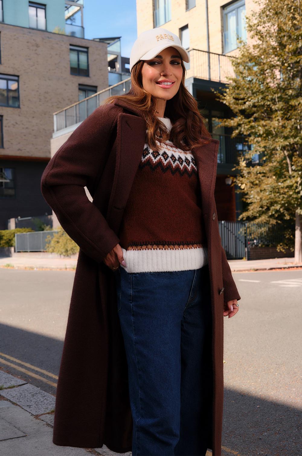 Personne portant un manteau en laine marron par-dessus un pull à motif jacquard avec un jean et une casquette blanche, dans un décor urbain.