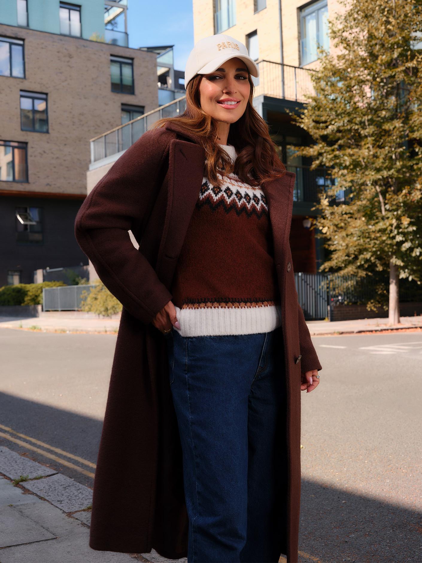 Brown wool coat worn over Fair Isle knit jumper with jeans and beige cap in urban setting.