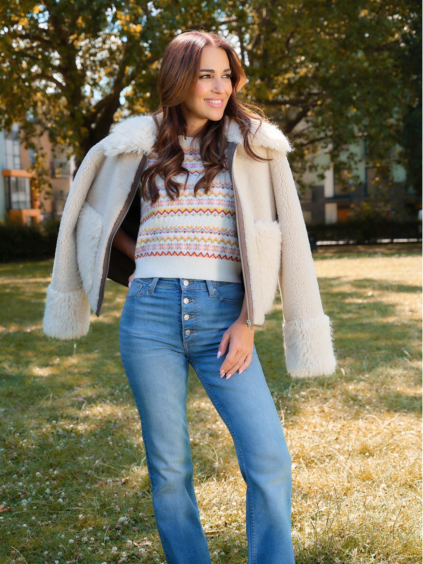 Cream shearling jacket worn over patterned knit jumper with blue button-front jeans in autumn outdoor setting.