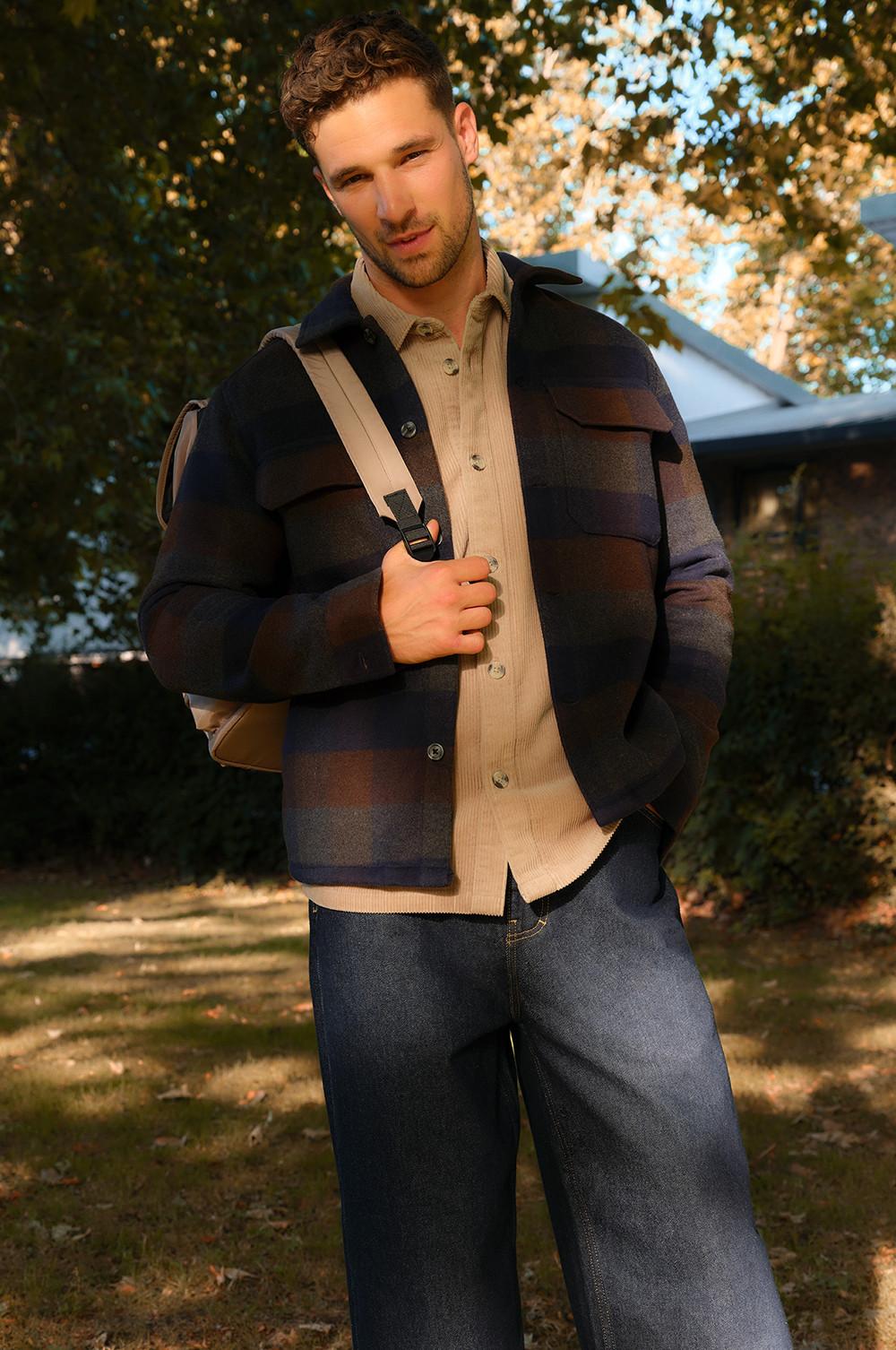 Person wearing navy checked overshirt, beige button-up shirt and denim jeans with shoulder bag outdoors.