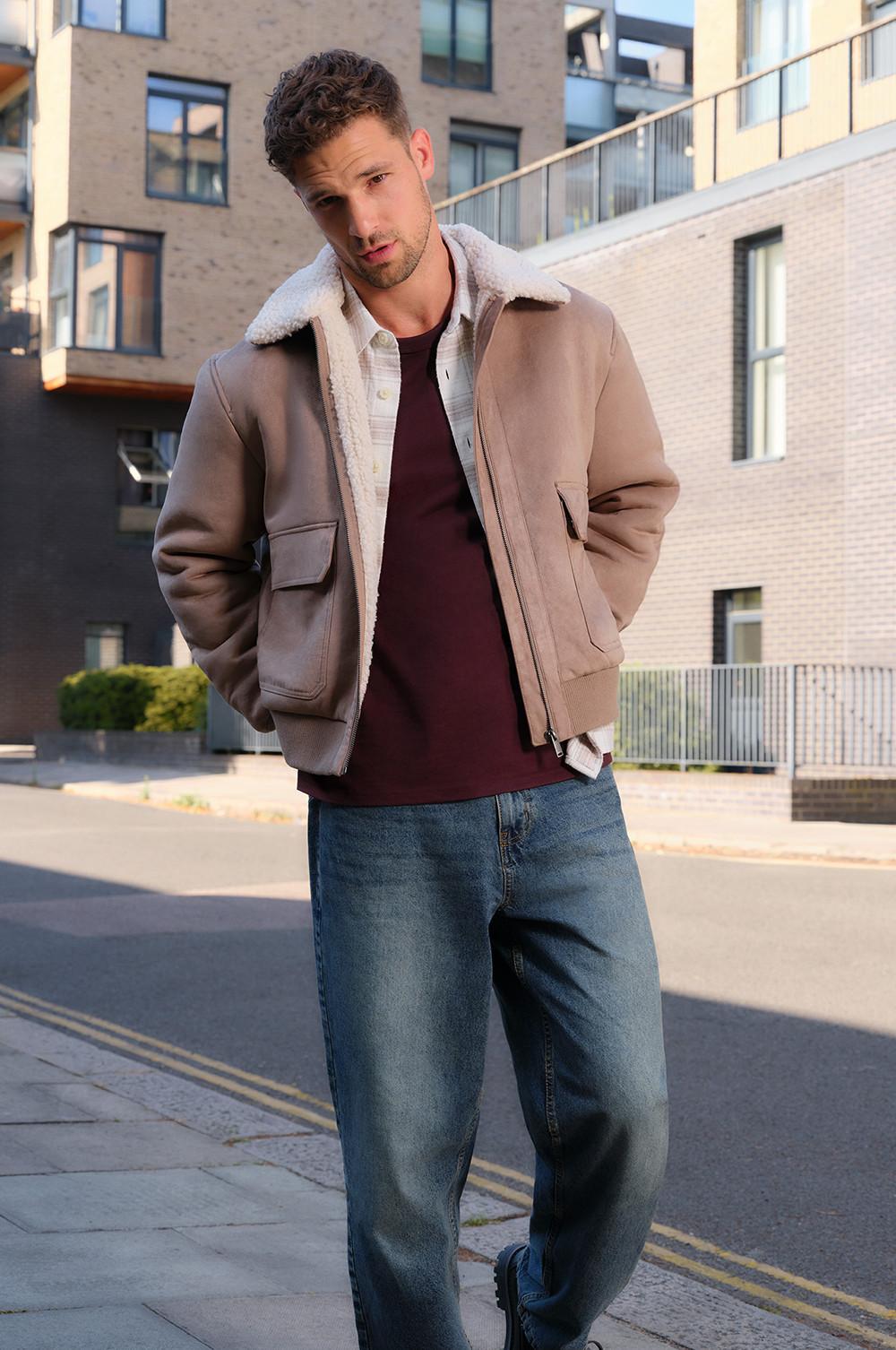 Person wearing beige shearling jacket over burgundy top and blue jeans, standing in urban setting.