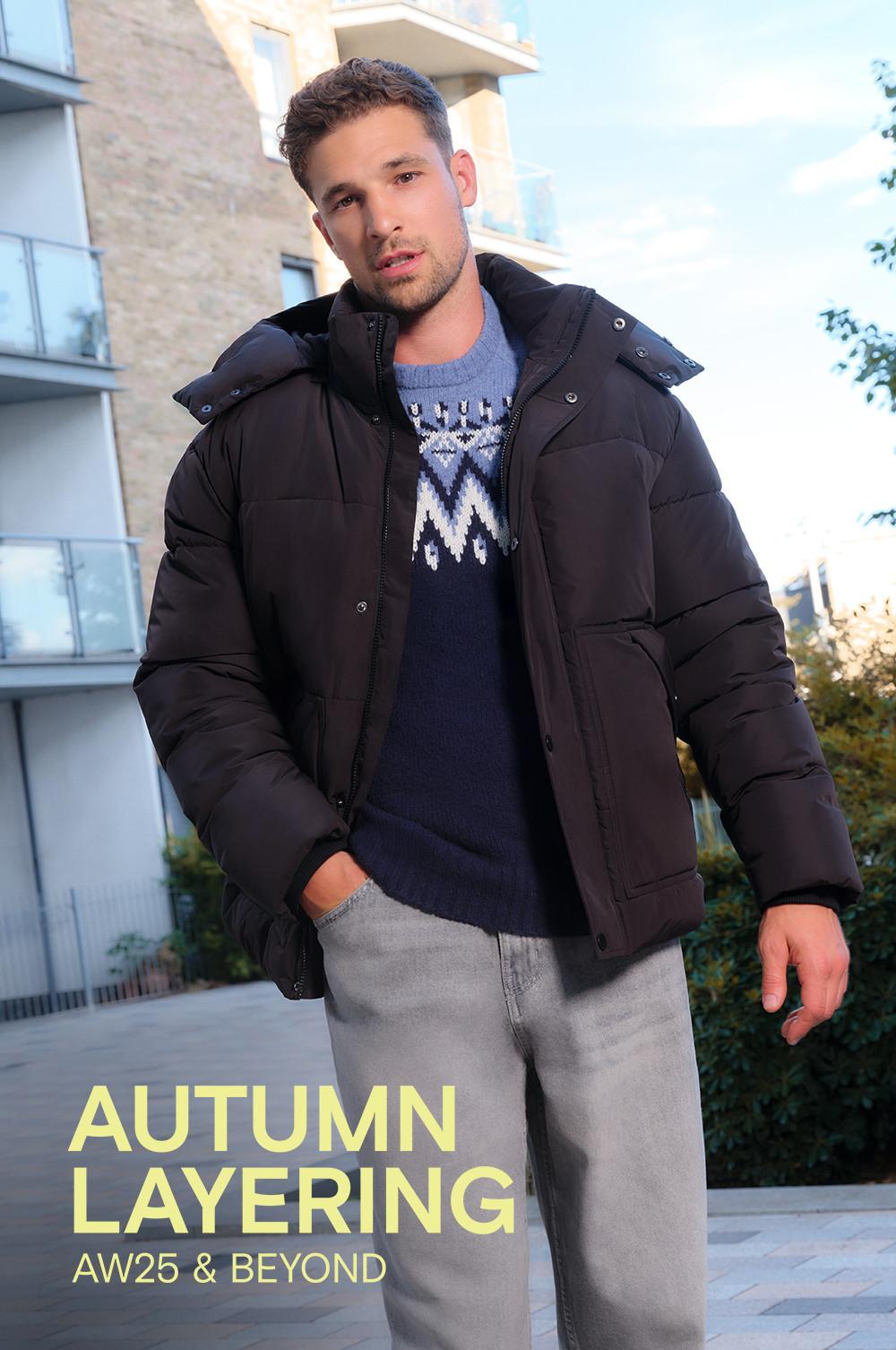 Person wearing black puffer jacket over patterned jumper with grey trousers outdoors. Text reads "AUTUMN LAYERING AW25 & BEYOND".
