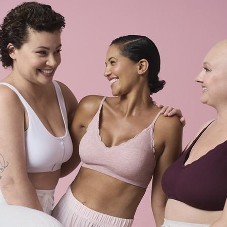 Three women wearing different coloured bras smiling at each other against a pink background.