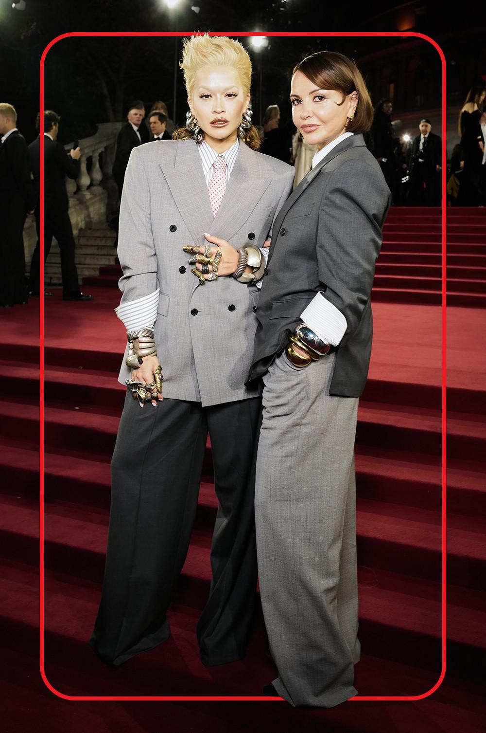 Rita Ora and Vera Ora wearing oversized grey blazers, wide leg trousers, white striped shirts and large silver statement jewellery standing on red carpet steps..