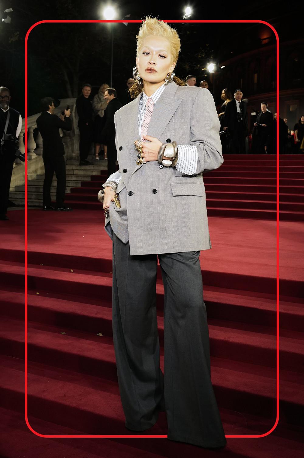 Rita Ora wearing grey double-breasted blazer with charcoal wide leg trousers, a buttoned up striped shirt with rolled up cuffs paired with a pink spotty tie, large statement silver earrings and bracelets and multiple rings in siver and gold, on red carpet steps.