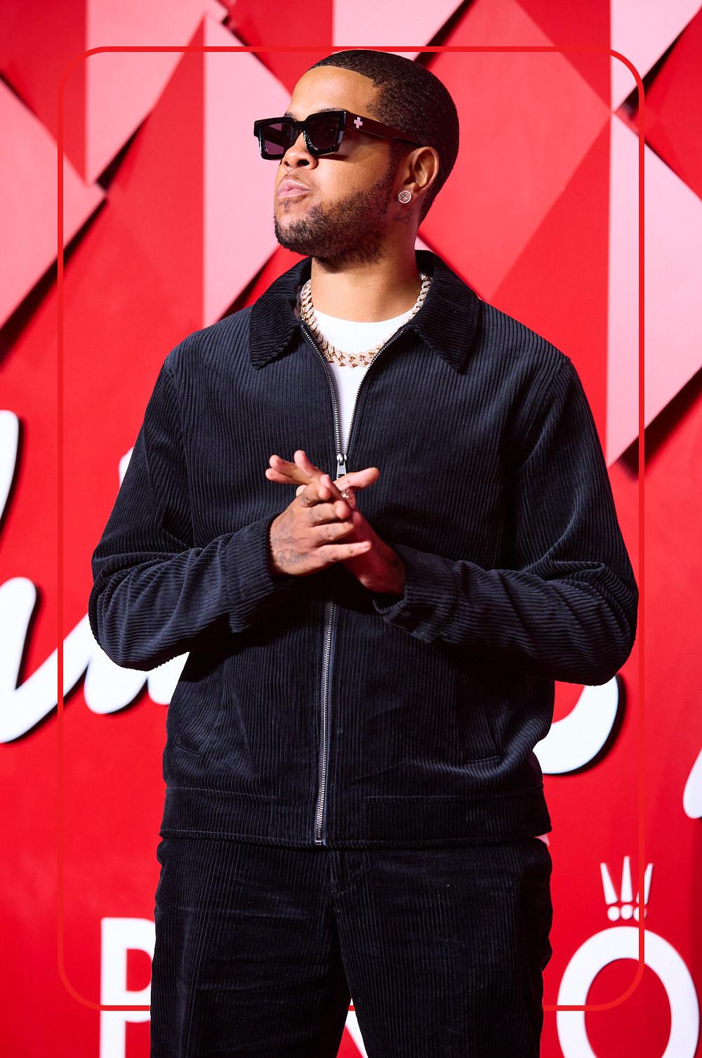 Chip wearing a navy corduroy jacket and matching trousers, white T-shirt, chain necklace and black square sunglasses against red geometric backdrop.