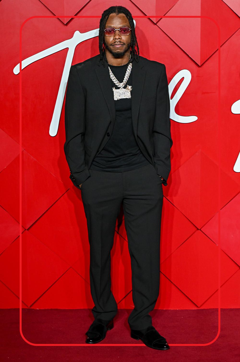 Krept black open blazer over black T-shirt with black smart trousers and black shoes. Paired with silver chain and red sunglasses against geometric red background.