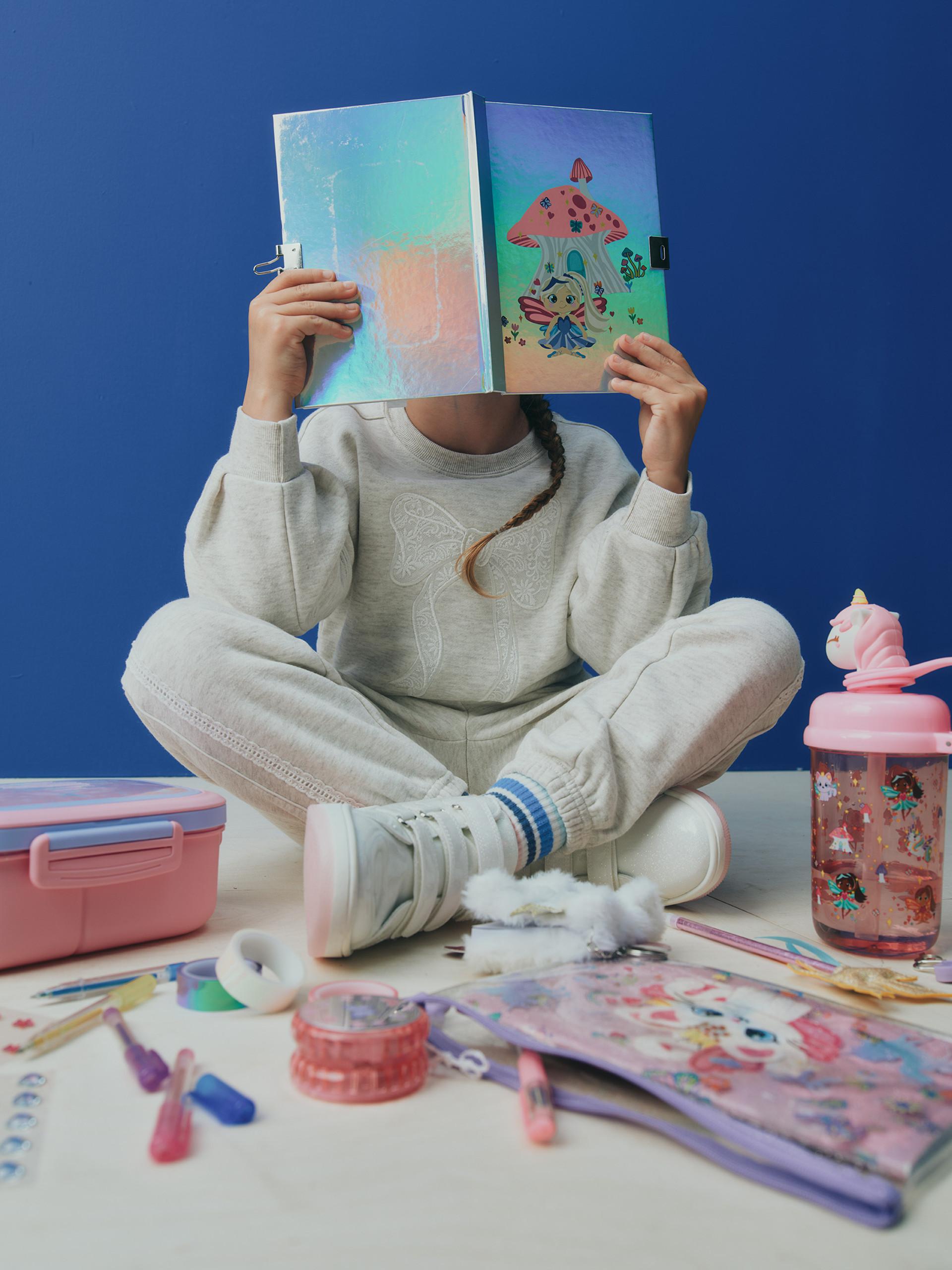 Model holding a book over their face wearing a grey loungewear set, sitting on the floor surrounded by stationary items