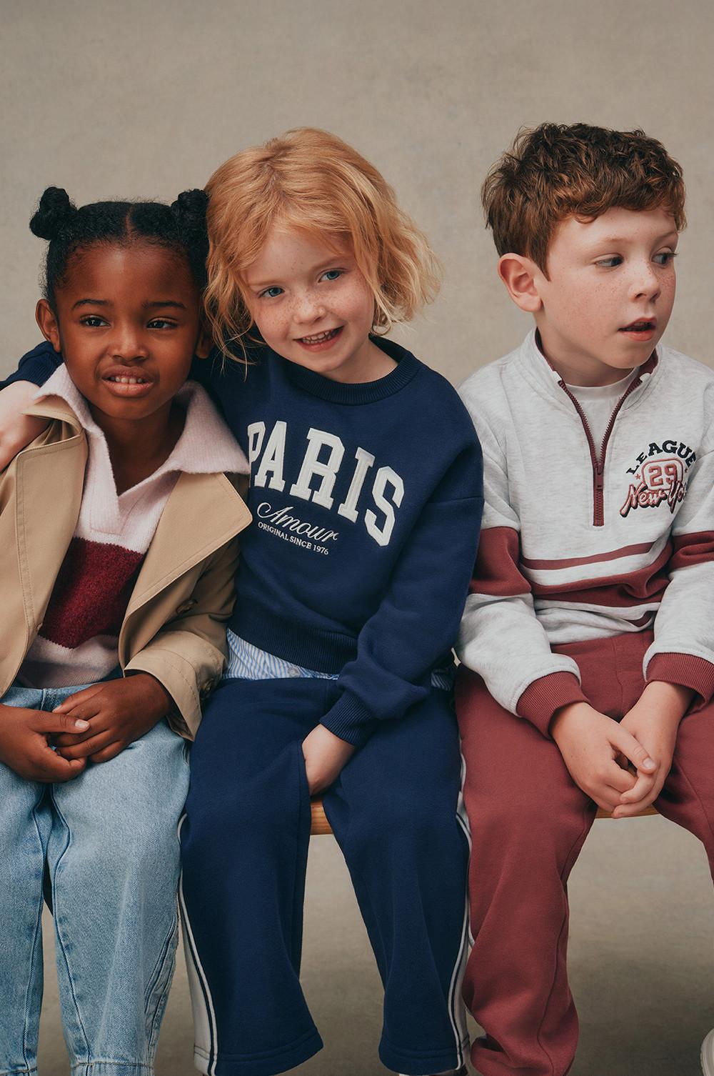 Three children wearing casual clothing including a blue PARIS sweatshirt, a trench coat, and a sporty pullover.