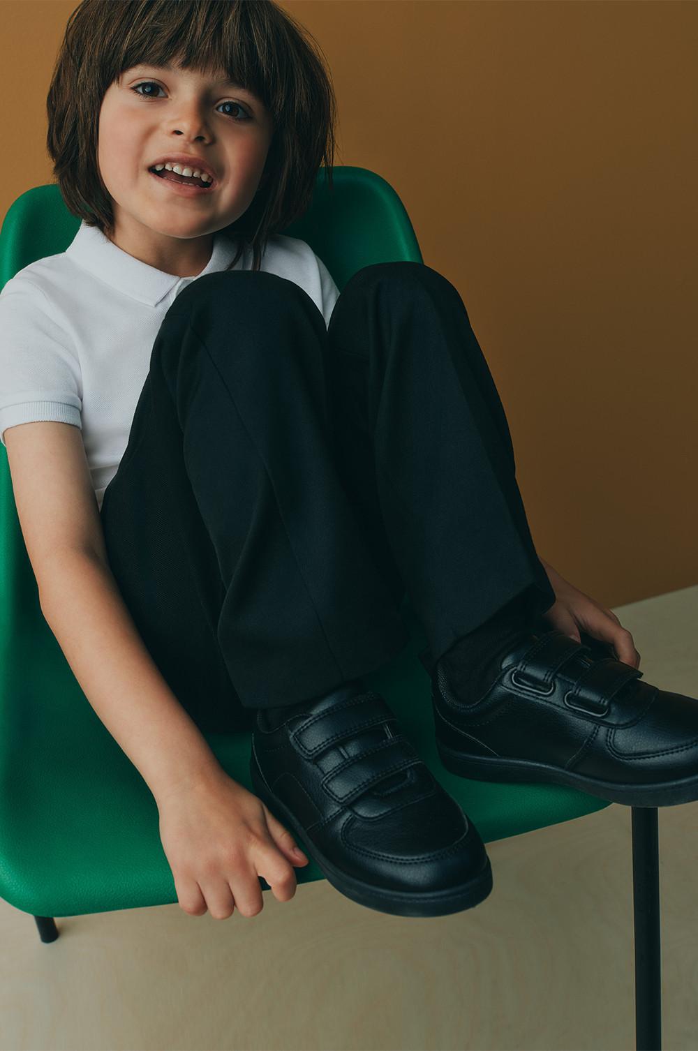 Child wearing school uniform with white polo shirt, black trousers and black shoes sitting in a green chair.