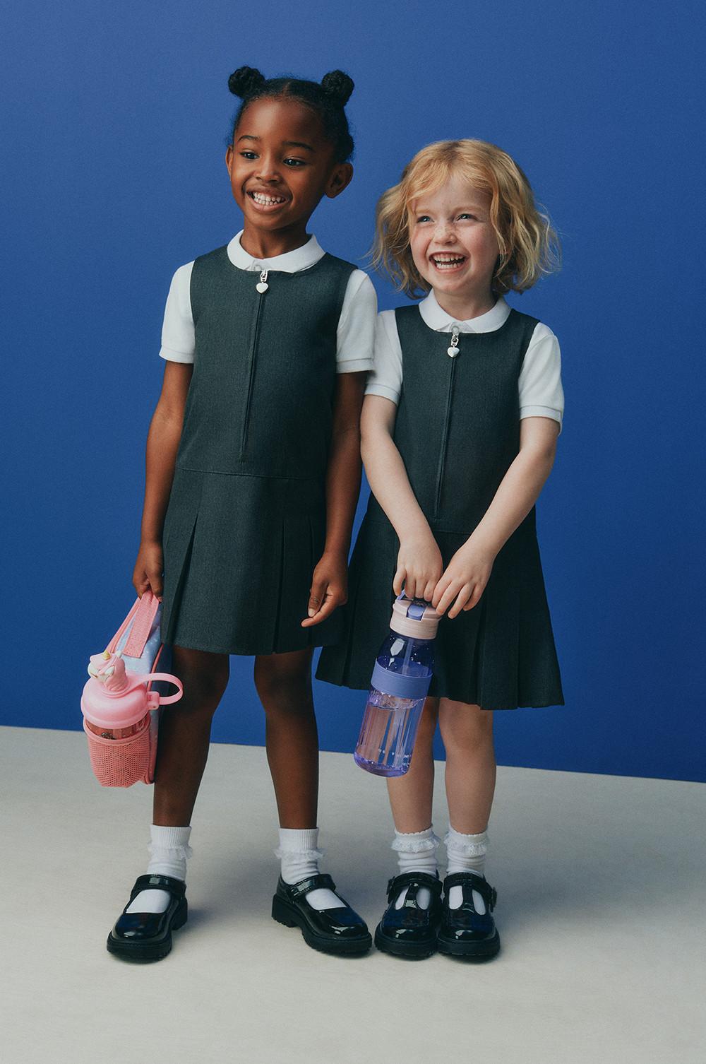 Two children wearing matching grey school pinafore dresses with white shirts, holding water bottles against a blue background.