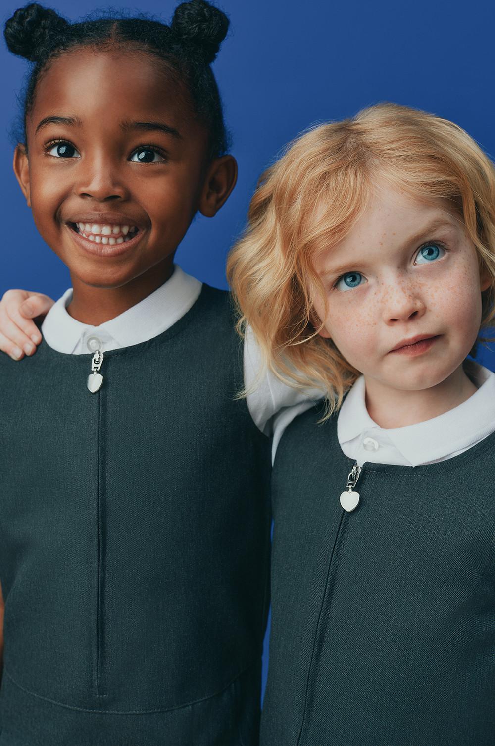 Two children wearing dark grey school uniform dressed with white collars and heart-shaped zipper pulls against a blue background.