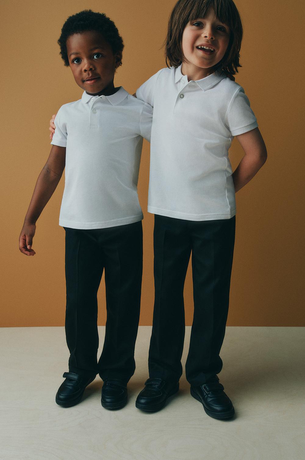 Two children wearing white polo shirts with dark trousers and black shoes standing together against a warm backdrop.